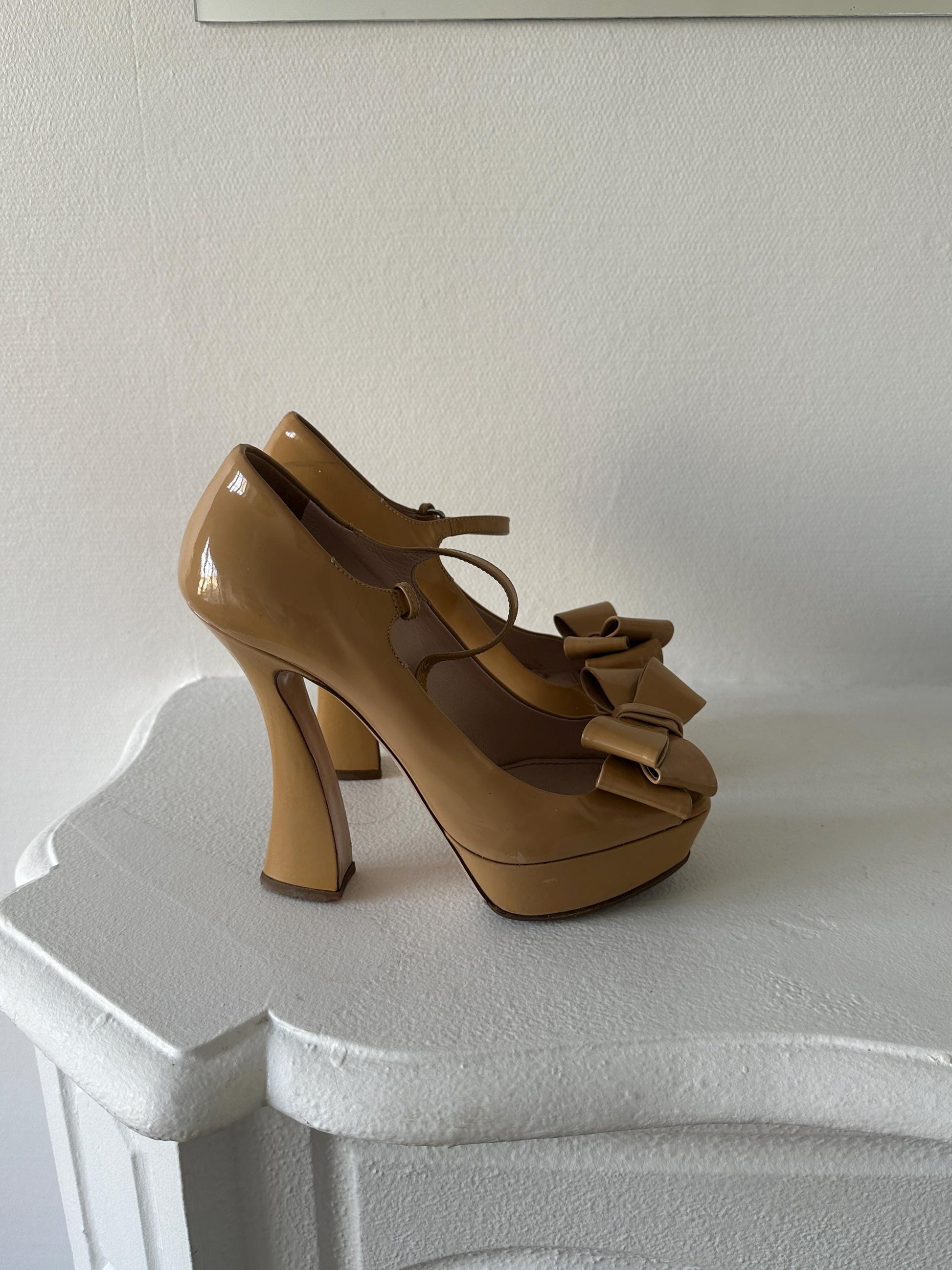 A pair of tan high-heeled shoes with bows on the toe are displayed against a white wall.