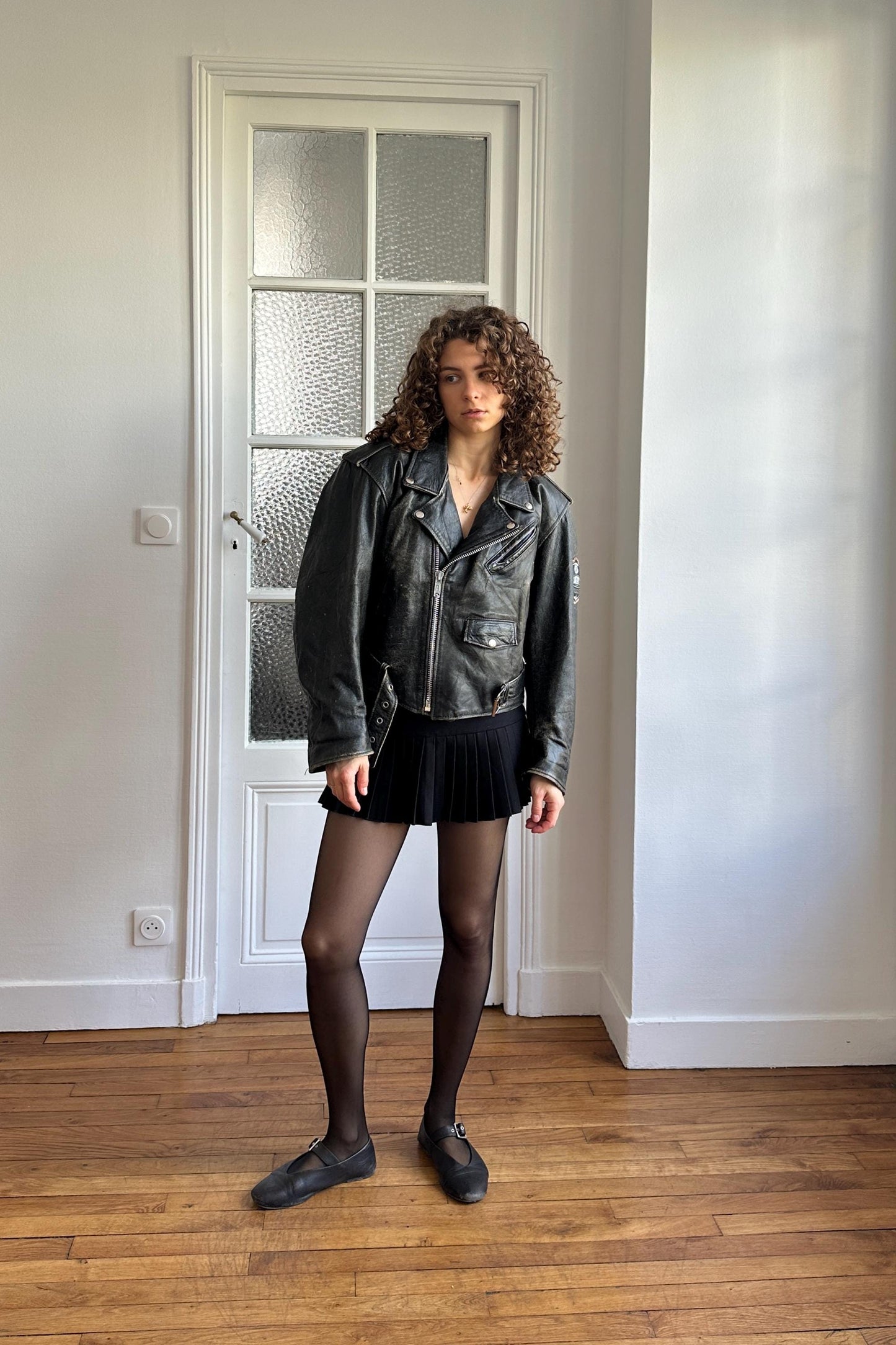 A woman in a black leather jacket and skirt stands in a room with a white door and wooden floor.