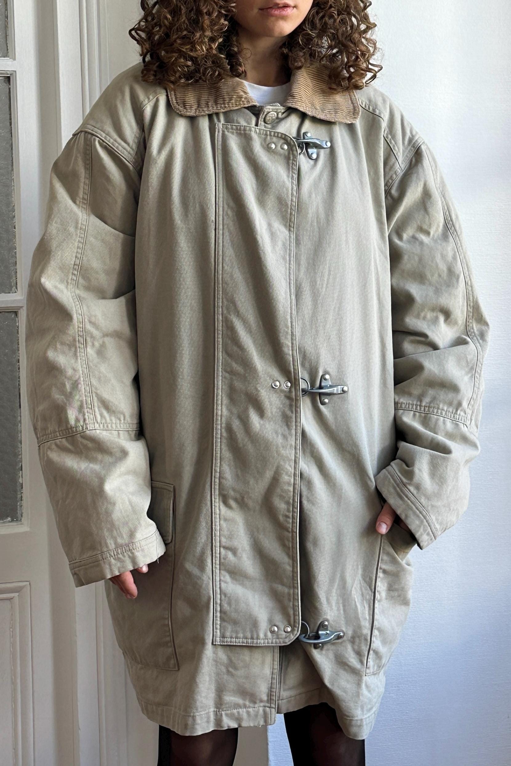 A person wearing a tan coat with a collar and metal buttons, standing in front of a white door.