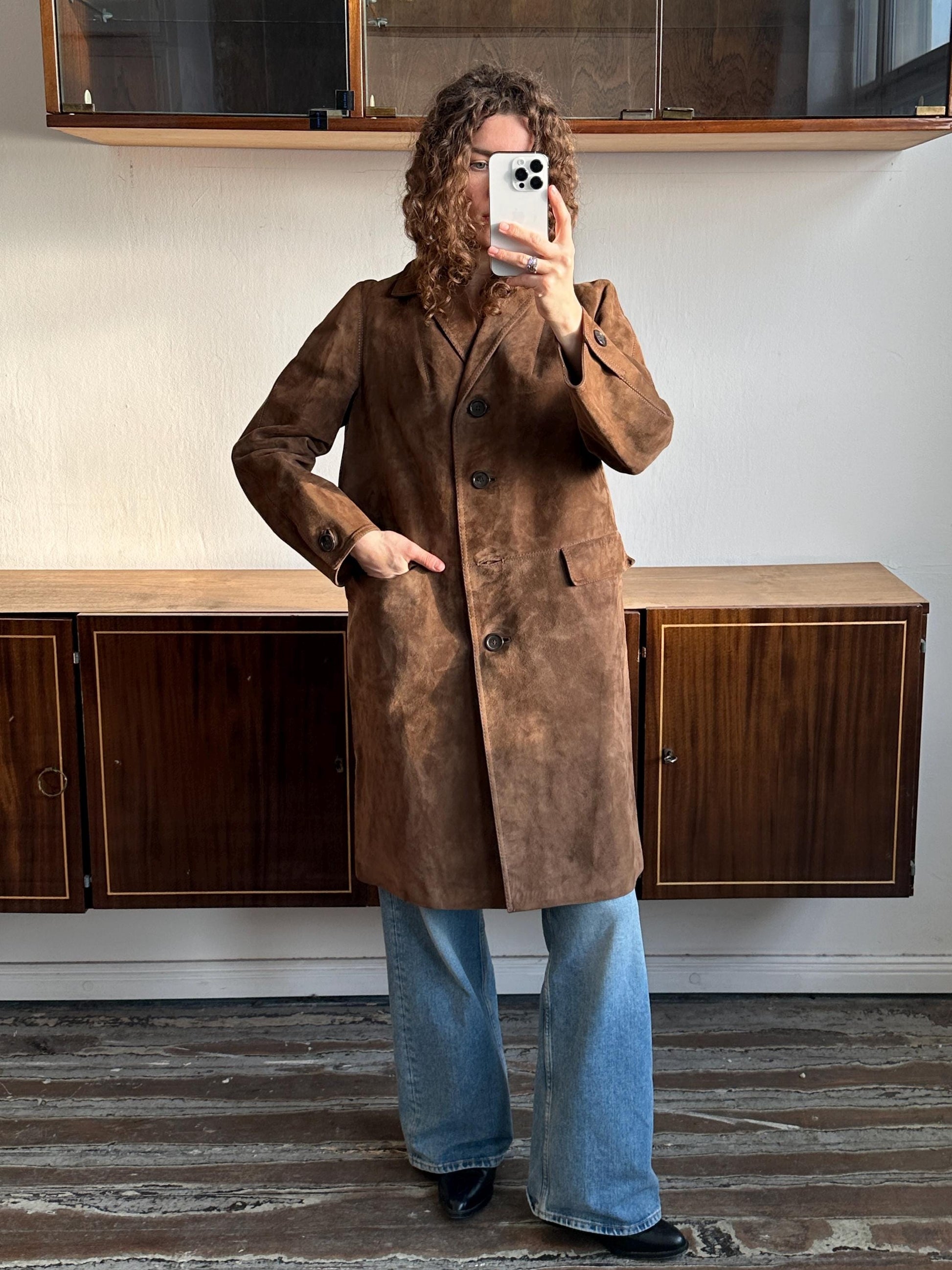 A person is taking a selfie with a brown suede coat and blue jeans, standing in front of a wooden cabinet.