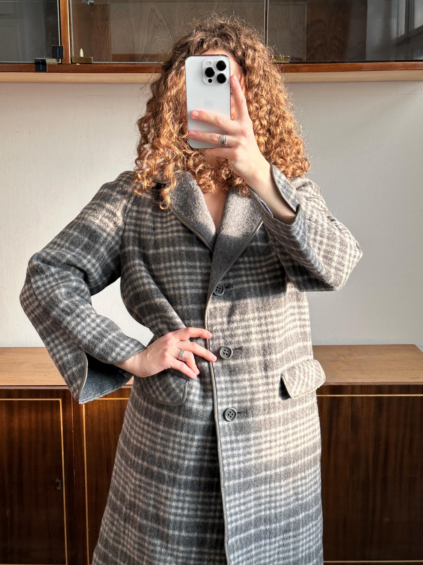 A person with curly hair is taking a selfie with a smartphone, wearing a gray and white plaid coat.