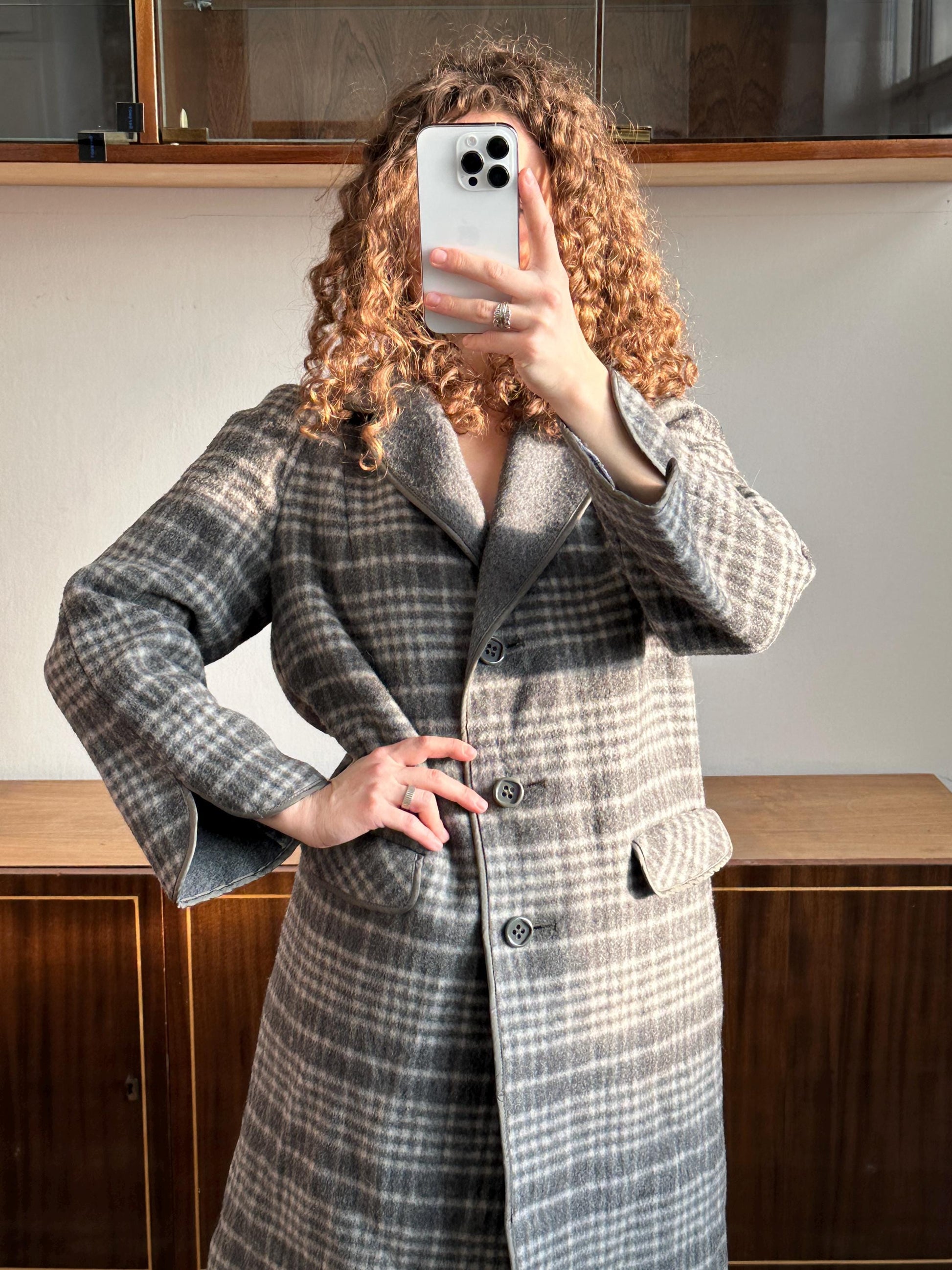A person with curly hair is taking a selfie with a smartphone, wearing a gray and white plaid coat.