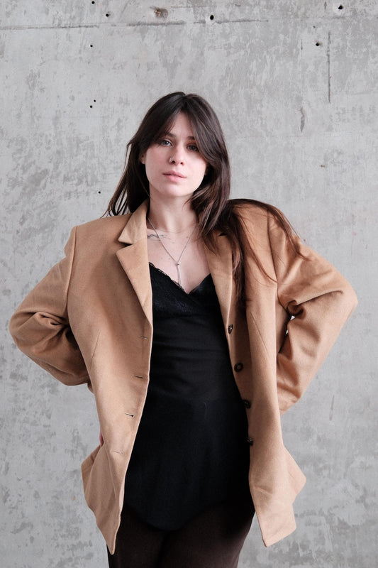 A woman wearing a brown jacket and black top is standing against a concrete wall.