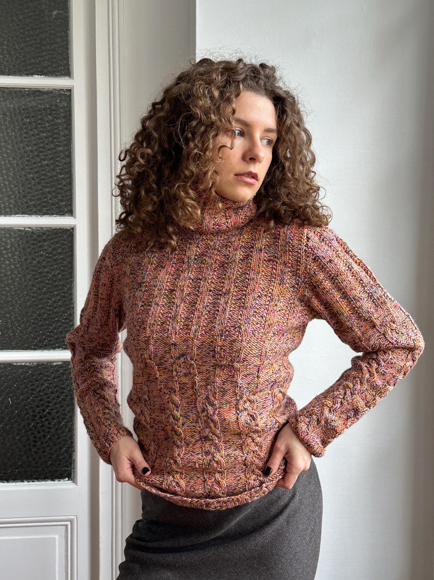 A woman with curly hair wearing a pink and orange patterned sweater is posing with her hands on her hips, looking off to the side.