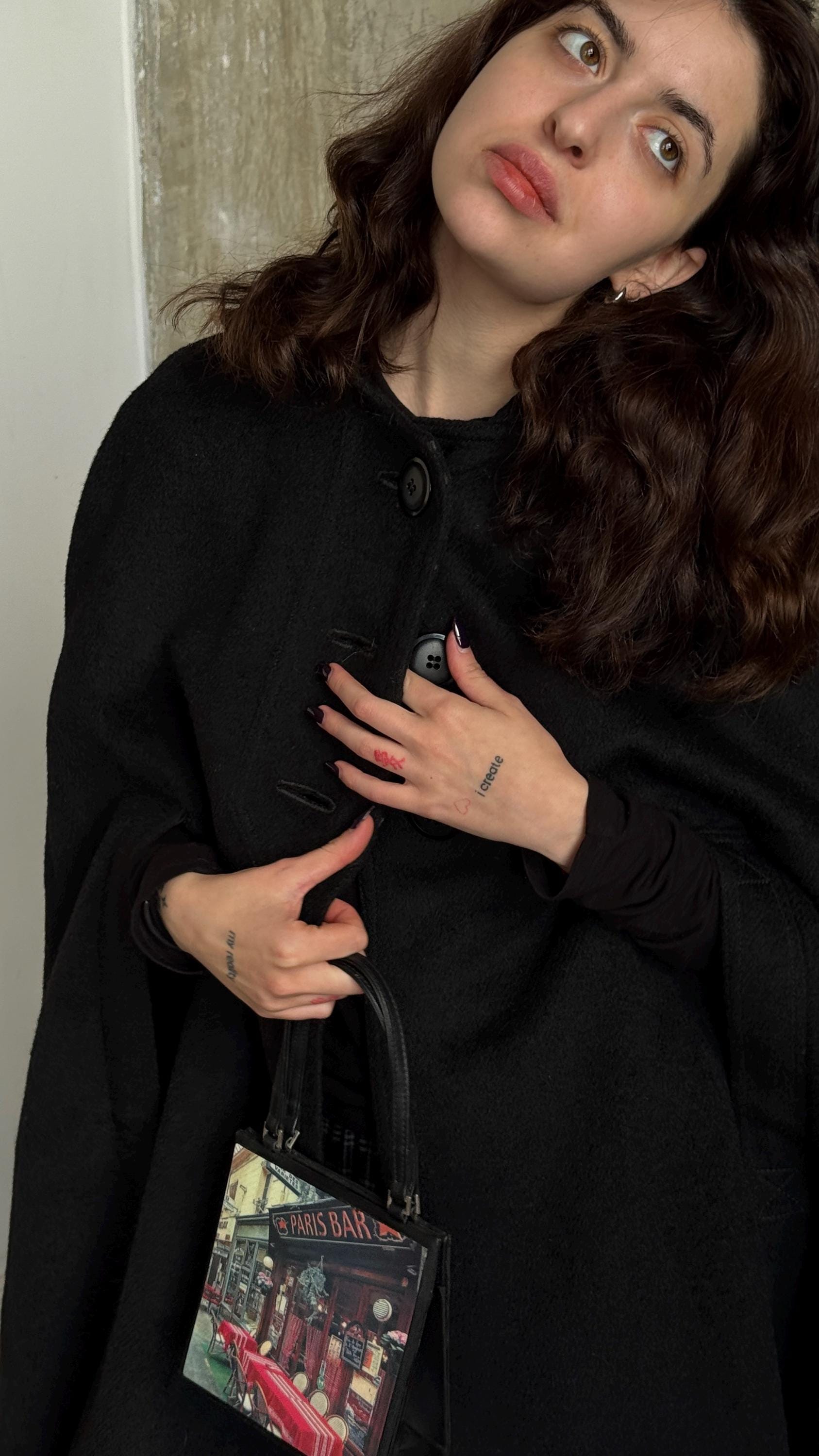 A woman with long brown hair wearing a black coat and carrying a Paris Bar bag stands in front of a beige wall.