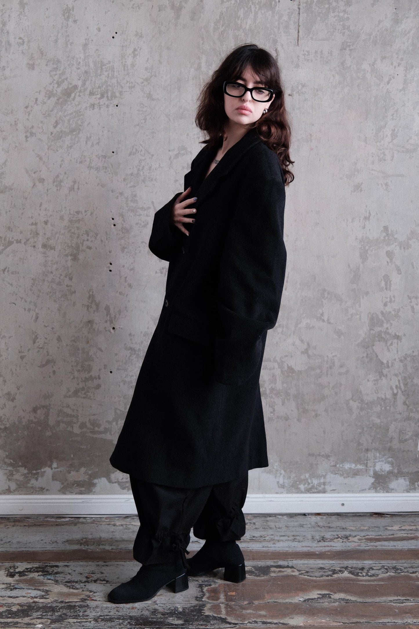 A woman in a long black coat stands against a plain gray wall, her gaze directed downwards.