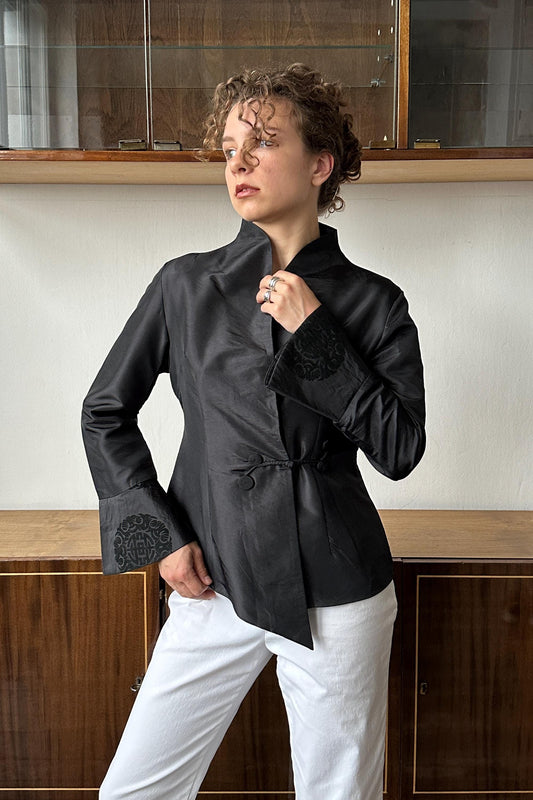 A woman wearing a black jacket with a unique design stands in front of a wooden cabinet.