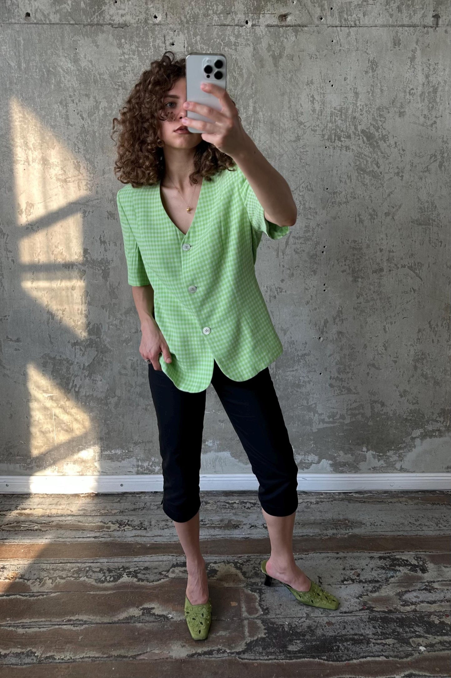 A woman with curly hair is taking a selfie with a smartphone, wearing a green and white checkered blazer and black pants, and green shoes.