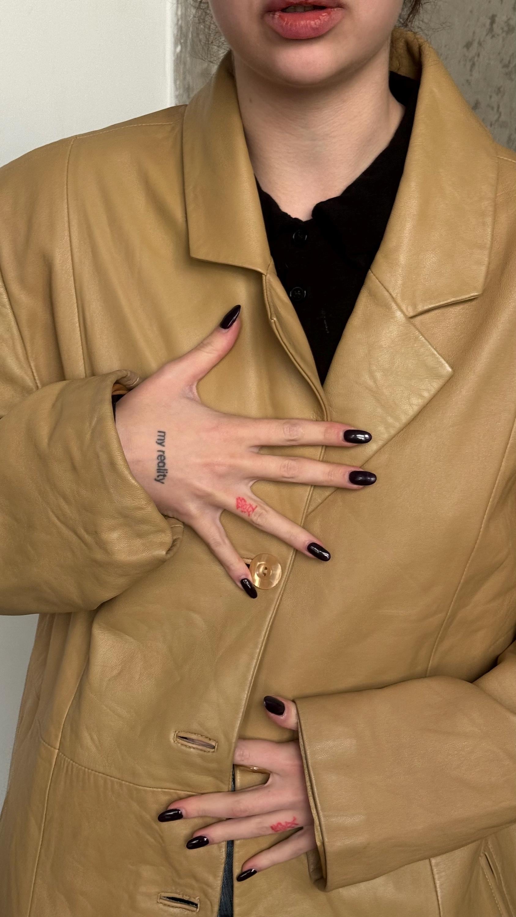 A close-up of a person wearing a tan leather jacket and holding their hands up to their chest.