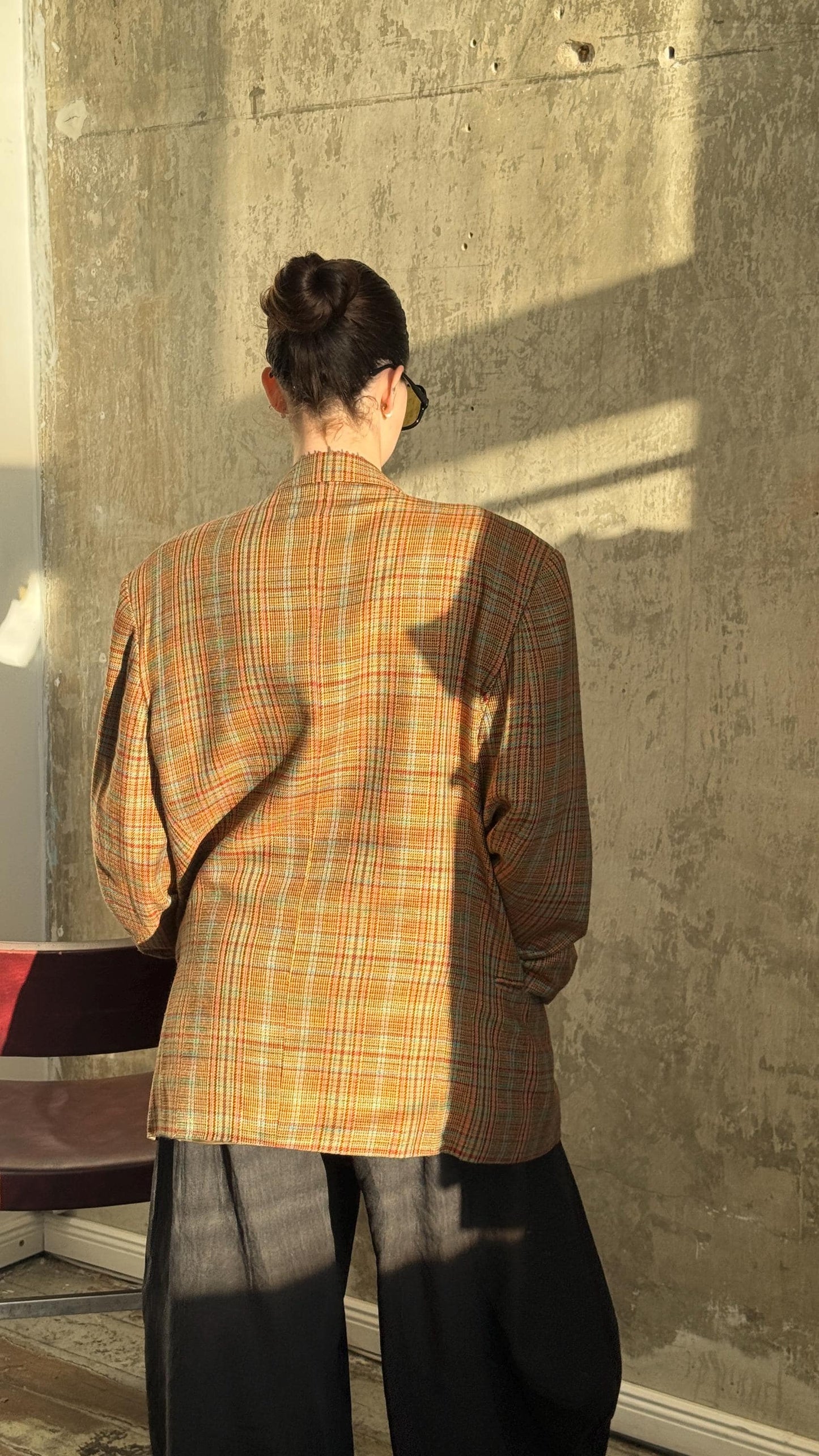 a person standing in front of a concrete wall, wearing a plaid jacket and sunglasses.