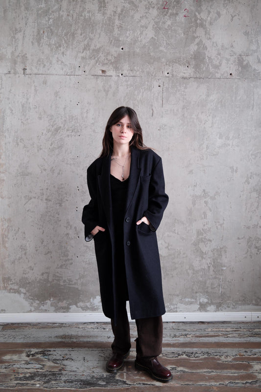 A woman in a long black coat stands in a room with a concrete wall.