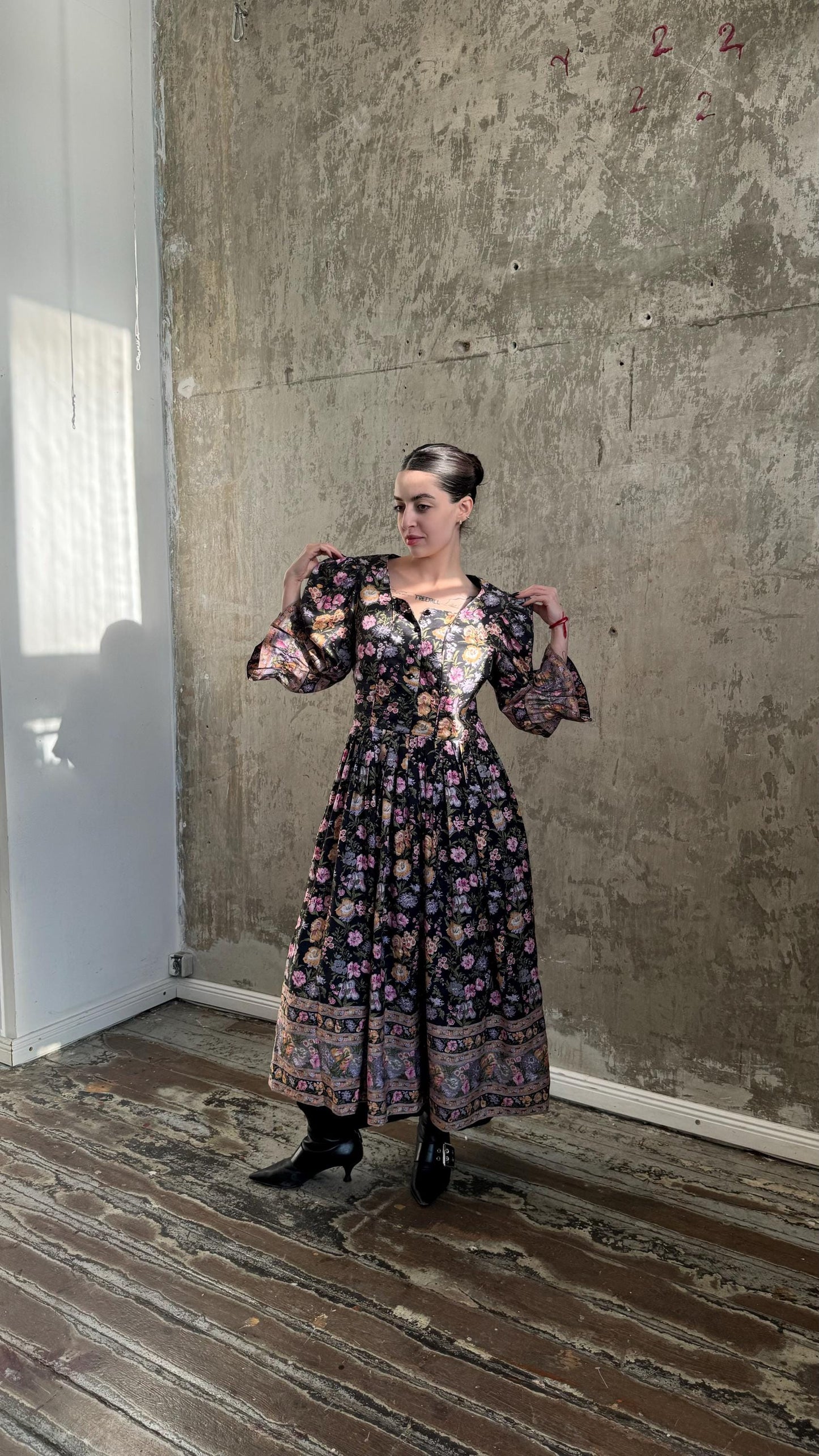 A woman wearing a long, floral-patterned dress stands in front of a concrete wall, with her hands behind her back.