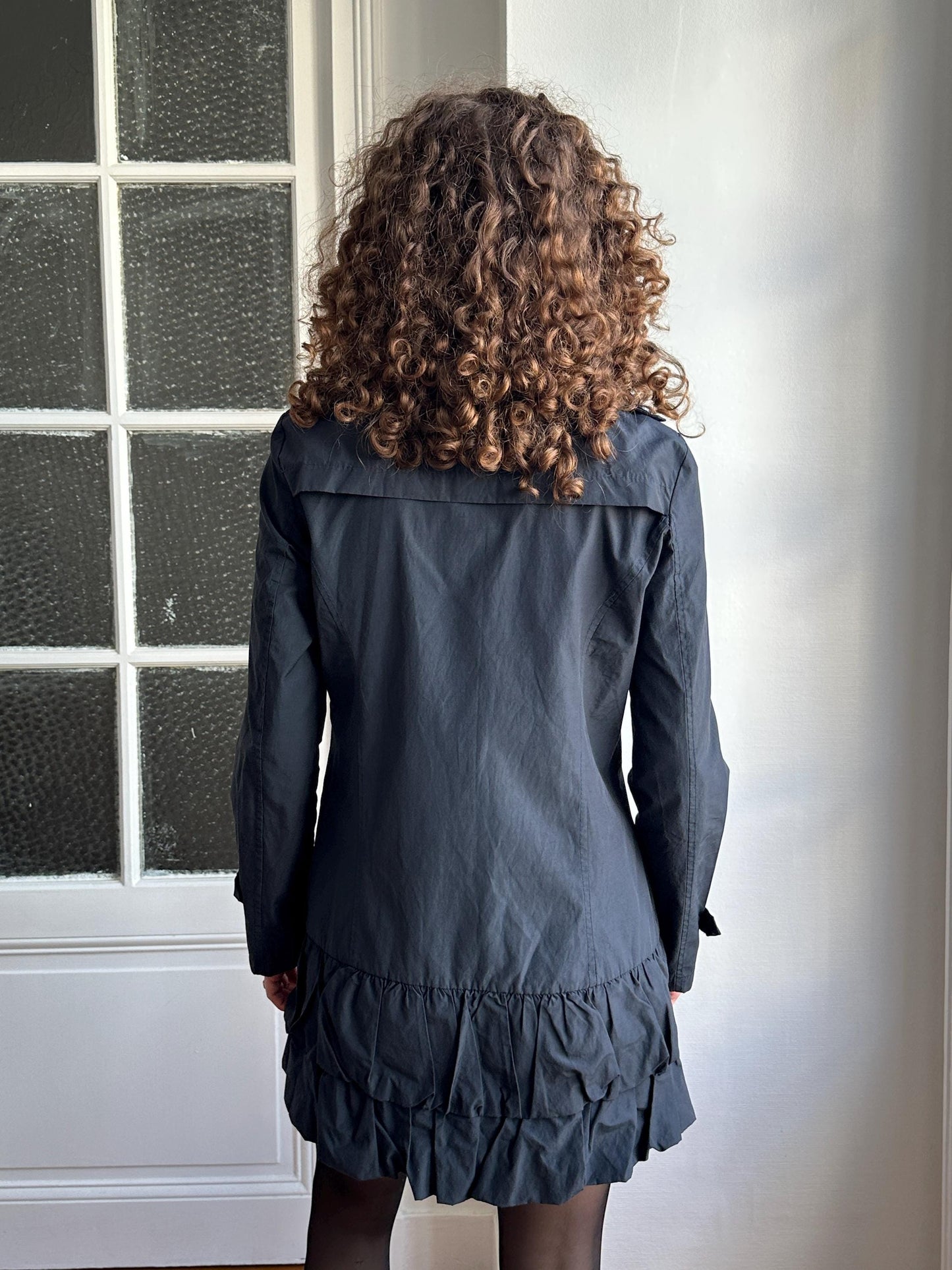 a person with curly hair wearing a black jacket and skirt, standing in front of a window.