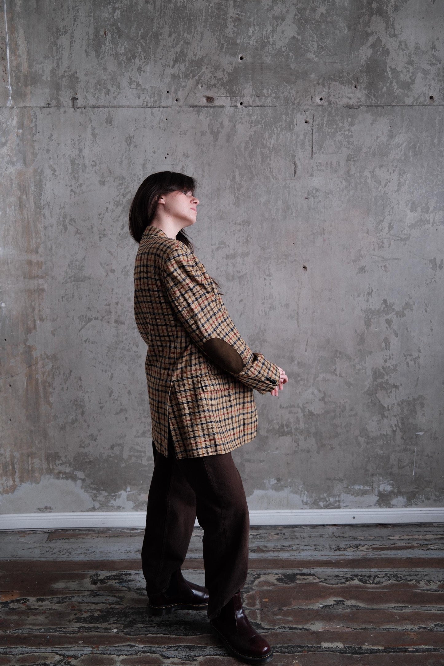 A person wearing a plaid jacket is standing in a room with a concrete wall.
