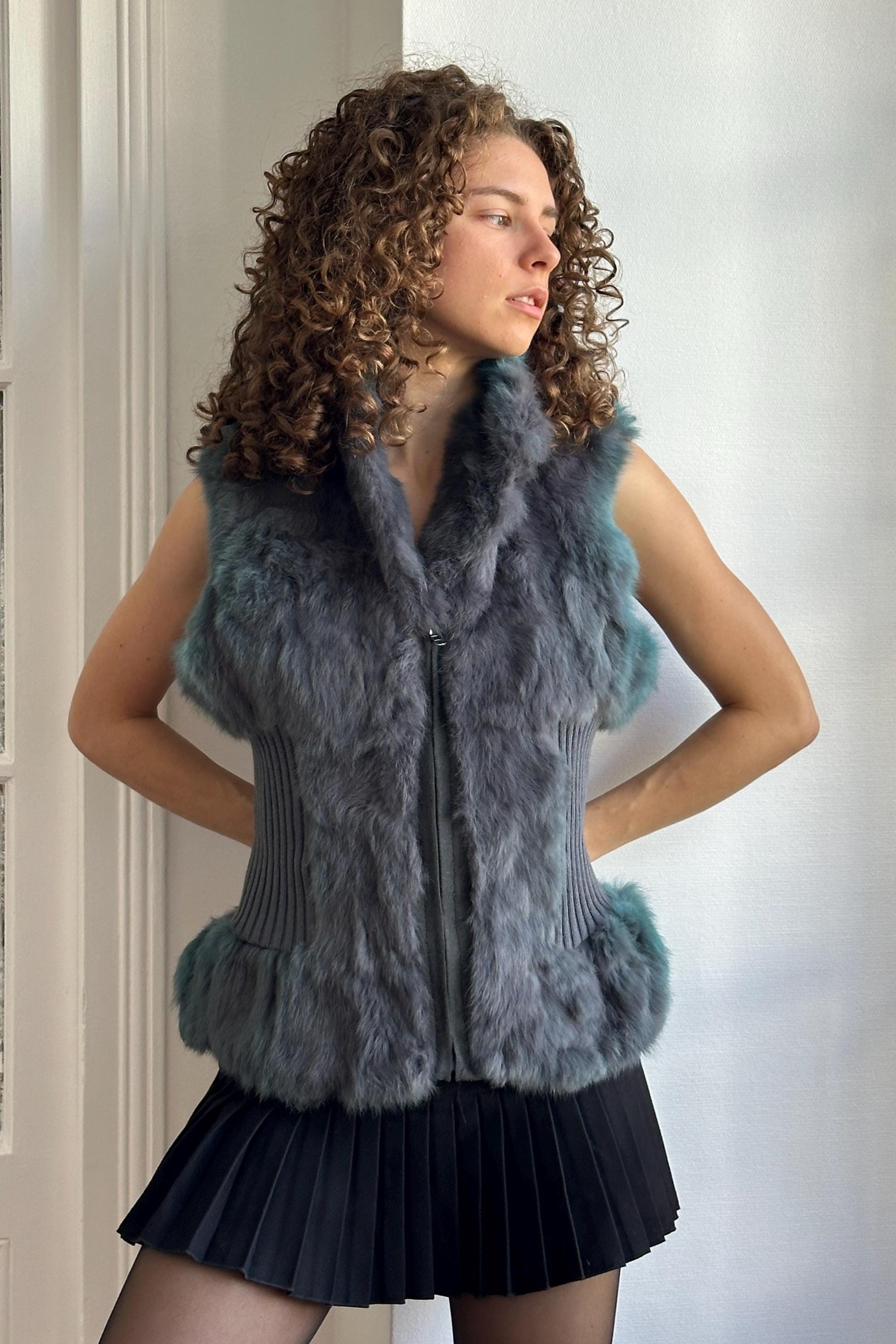 A woman with curly hair wearing a gray fur vest and a black skirt stands in front of a white wall.