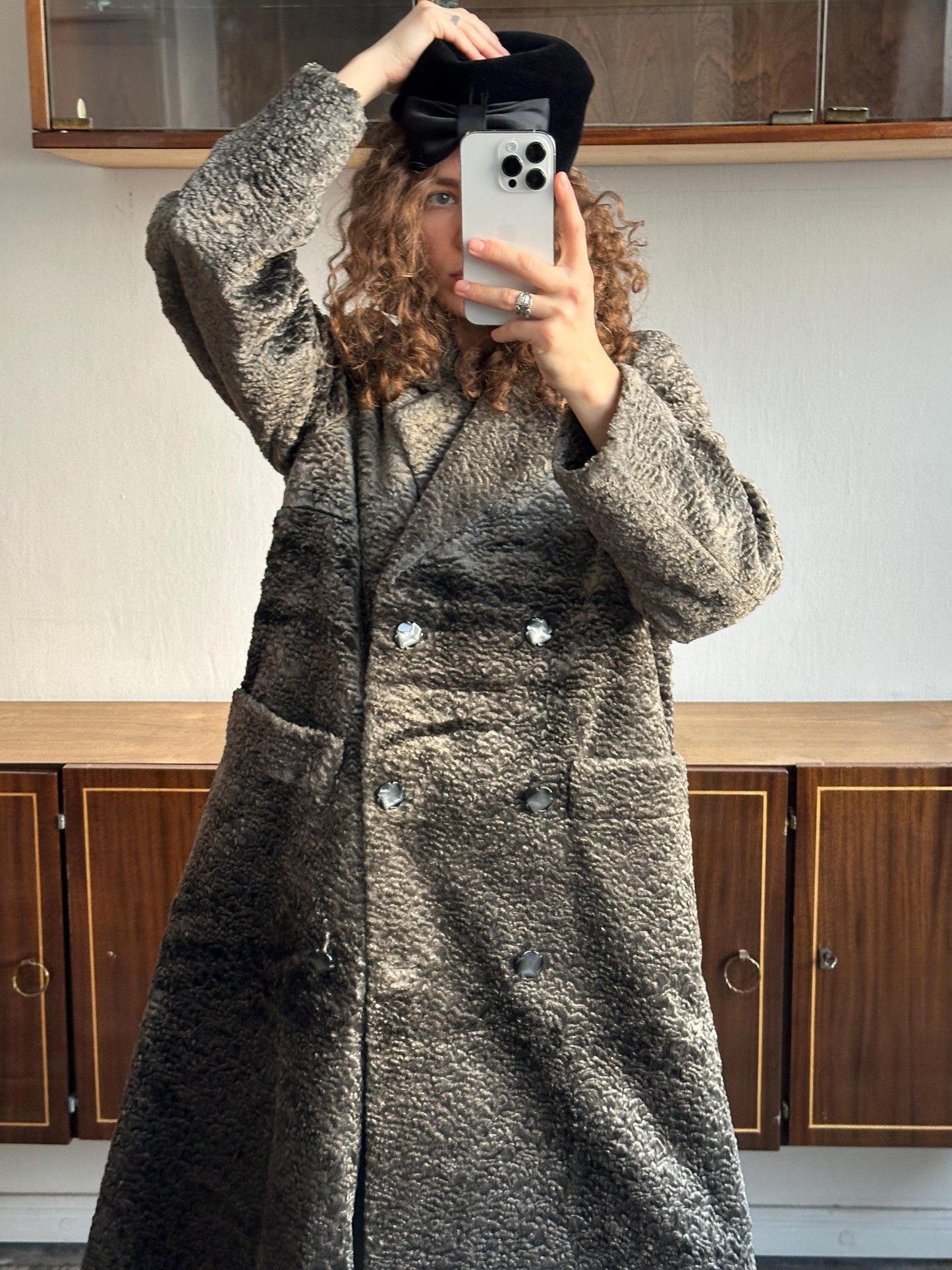A person taking a selfie with a black hat and a gray coat, standing in front of a wooden cabinet.