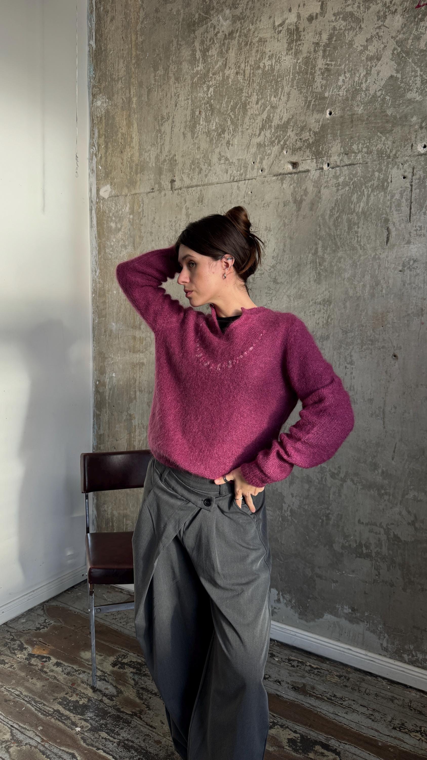 A woman wearing a pink sweater and gray pants stands in a room with a concrete wall and wooden floor.