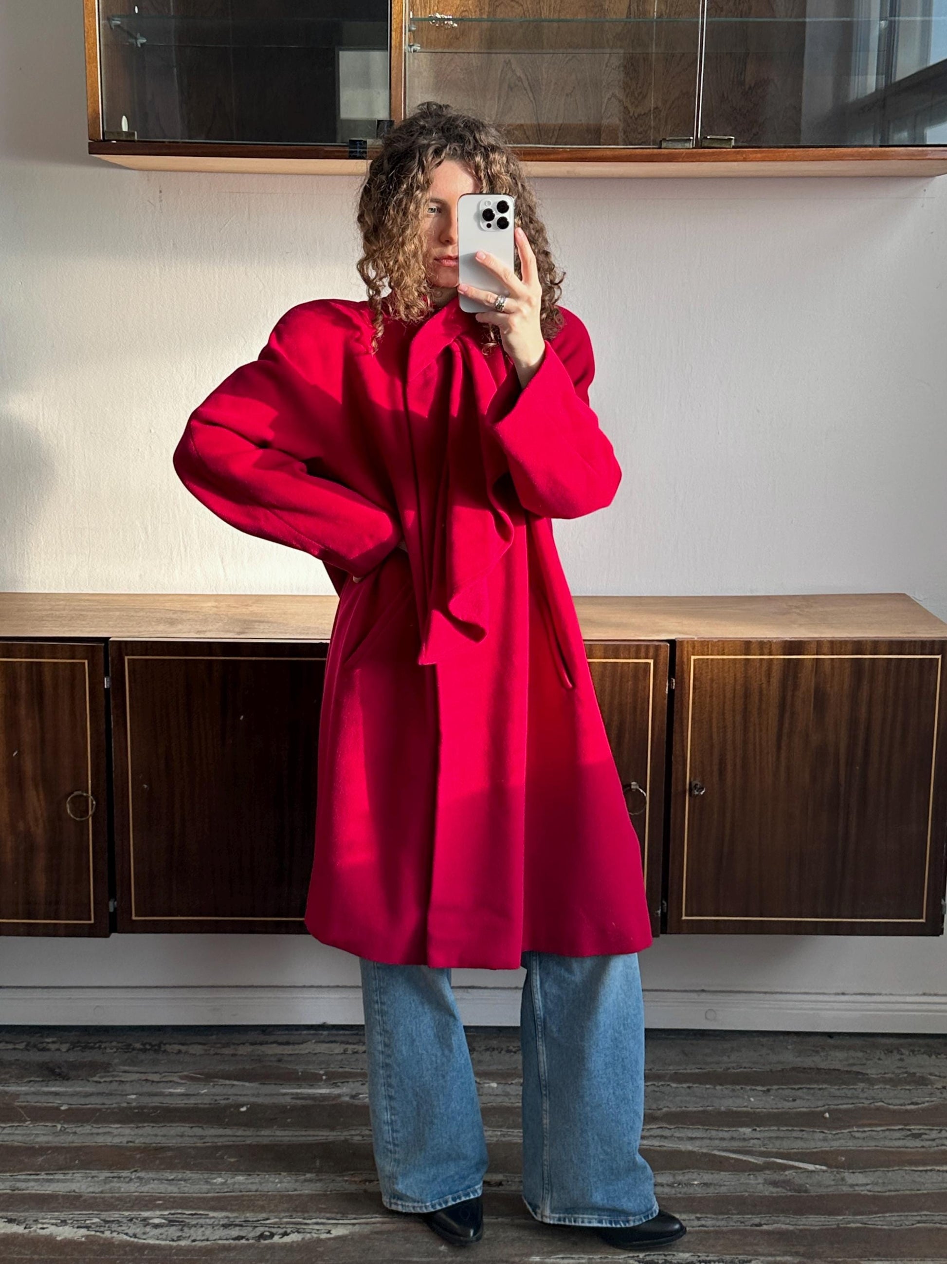 A person wearing a bright red coat and jeans is taking a selfie with a smartphone.