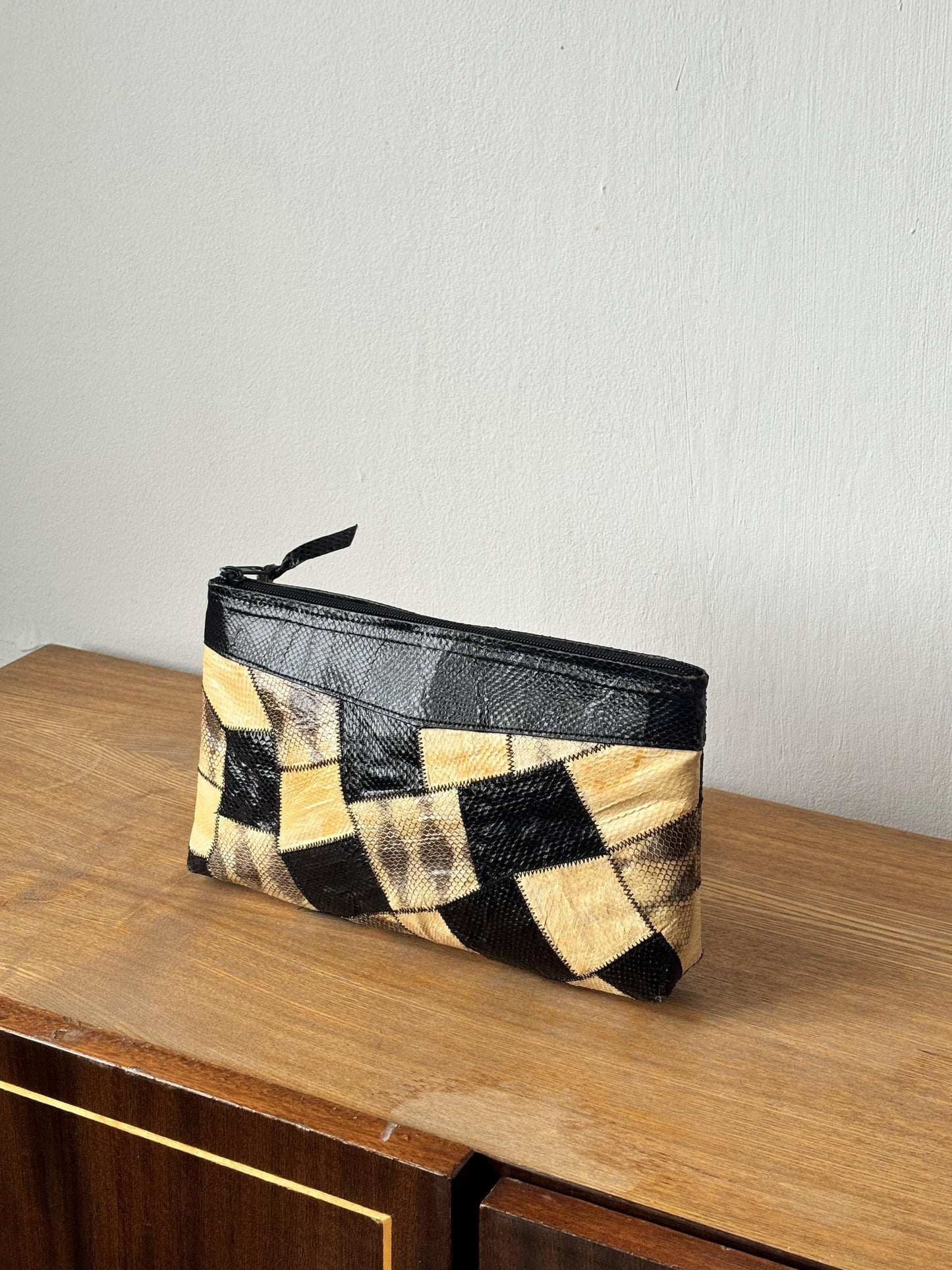 A black and beige patterned purse is sitting on a wooden surface against a white wall.