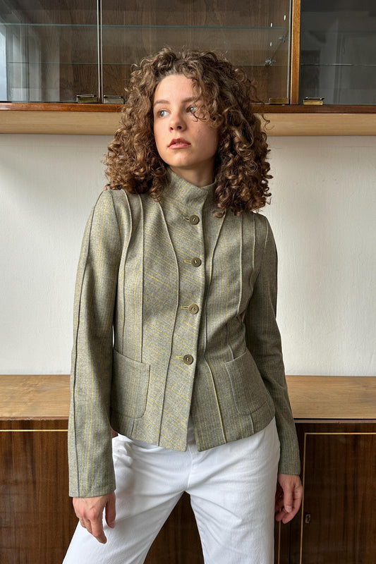 A woman with curly hair wearing a gray jacket and white pants stands in front of a wooden cabinet.