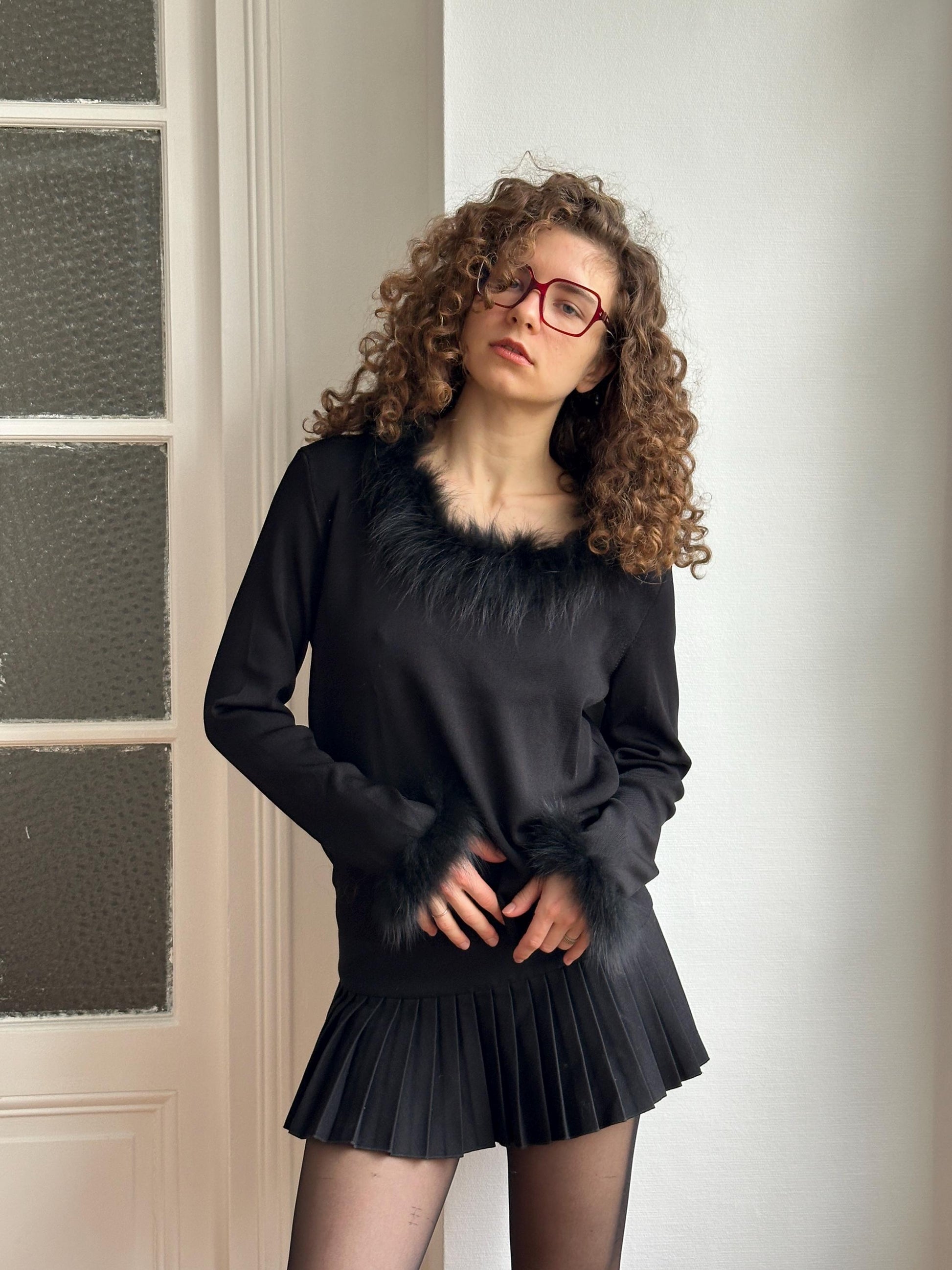 A woman with curly hair and glasses is posing in a black outfit with a fur collar, standing in front of a white door.