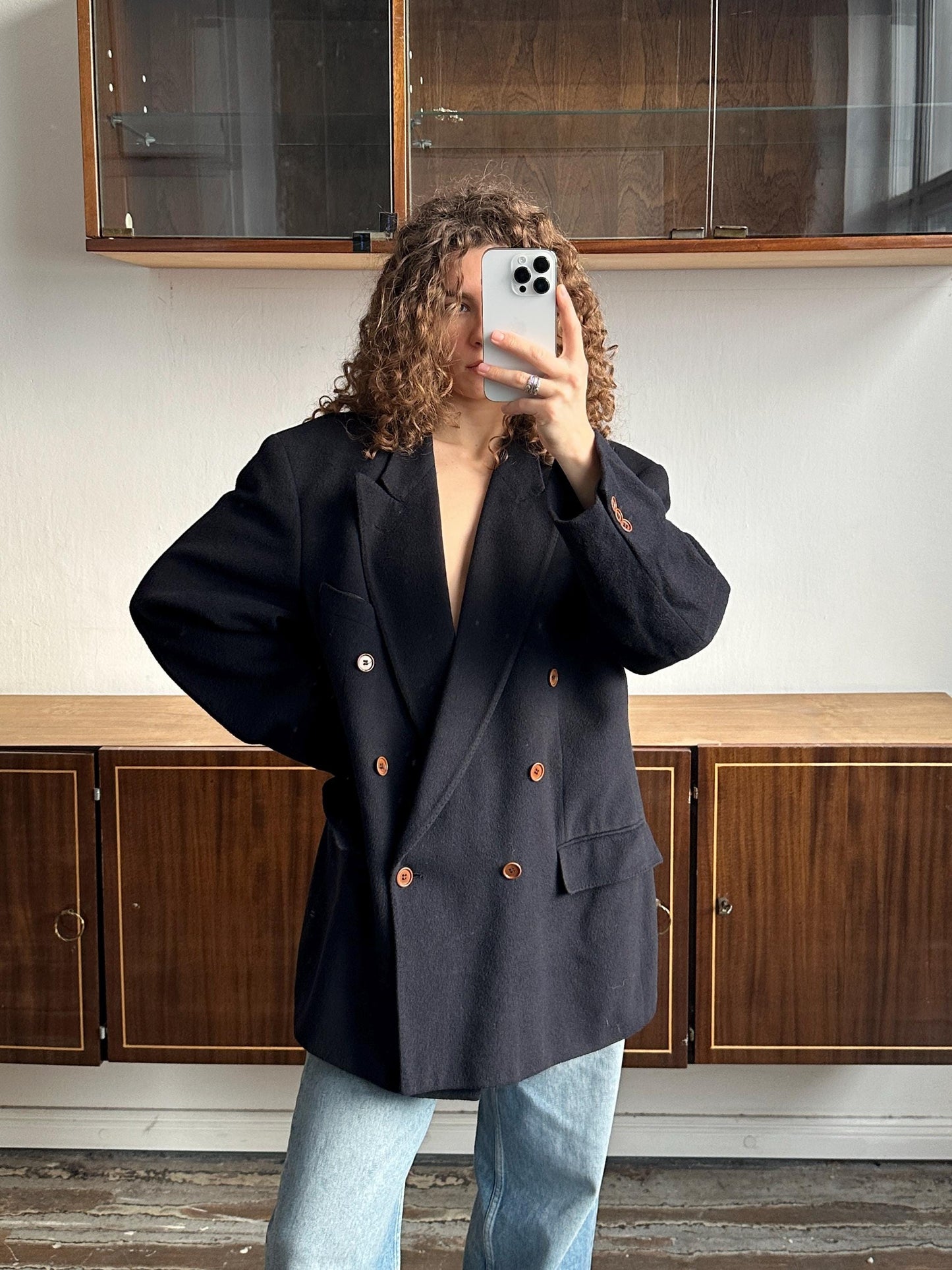 A person with curly hair is taking a selfie with a smartphone, wearing a black blazer and jeans.