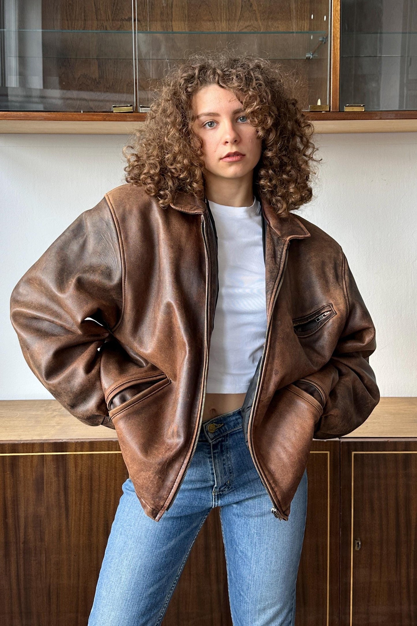A young woman with curly hair wearing a brown leather jacket and blue jeans stands in front of a wooden cabinet.