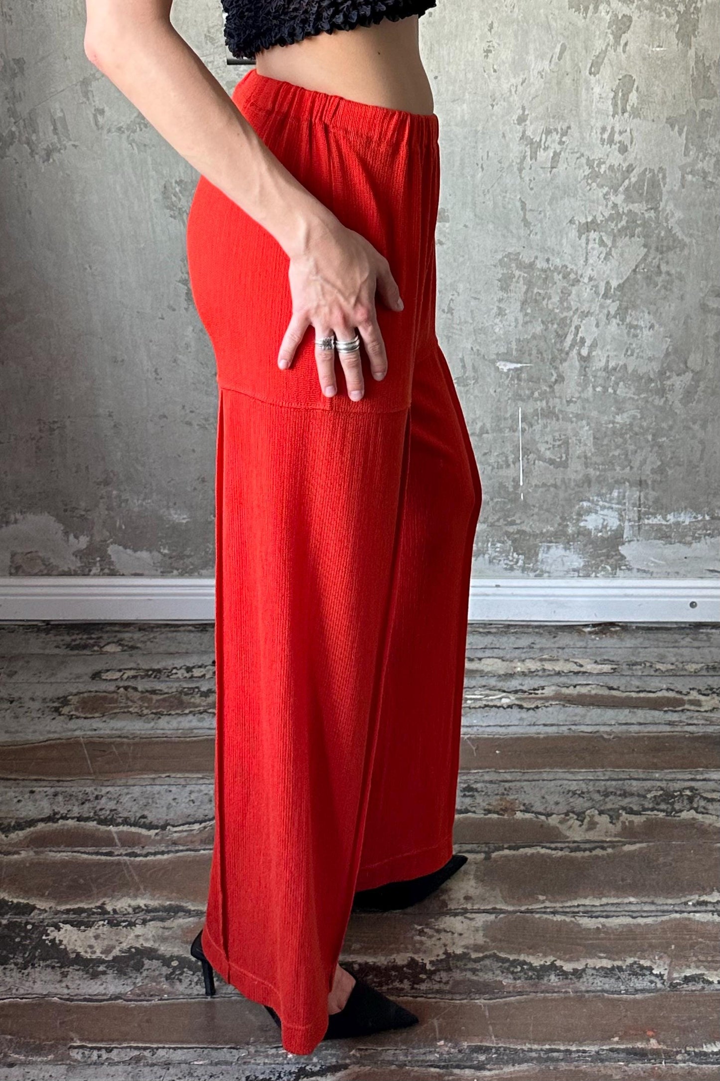 A person wearing a black top and red wide-leg pants stands in front of a concrete wall.