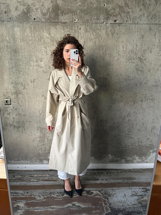 A woman in a beige trench coat is taking a selfie in front of a concrete wall.
