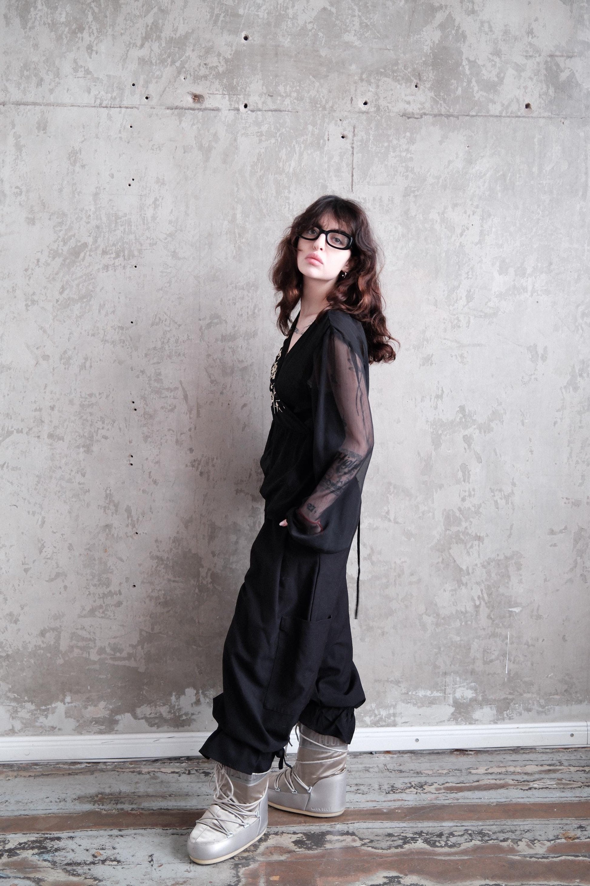 A woman in a black outfit stands against a concrete wall, her gaze directed downwards.
