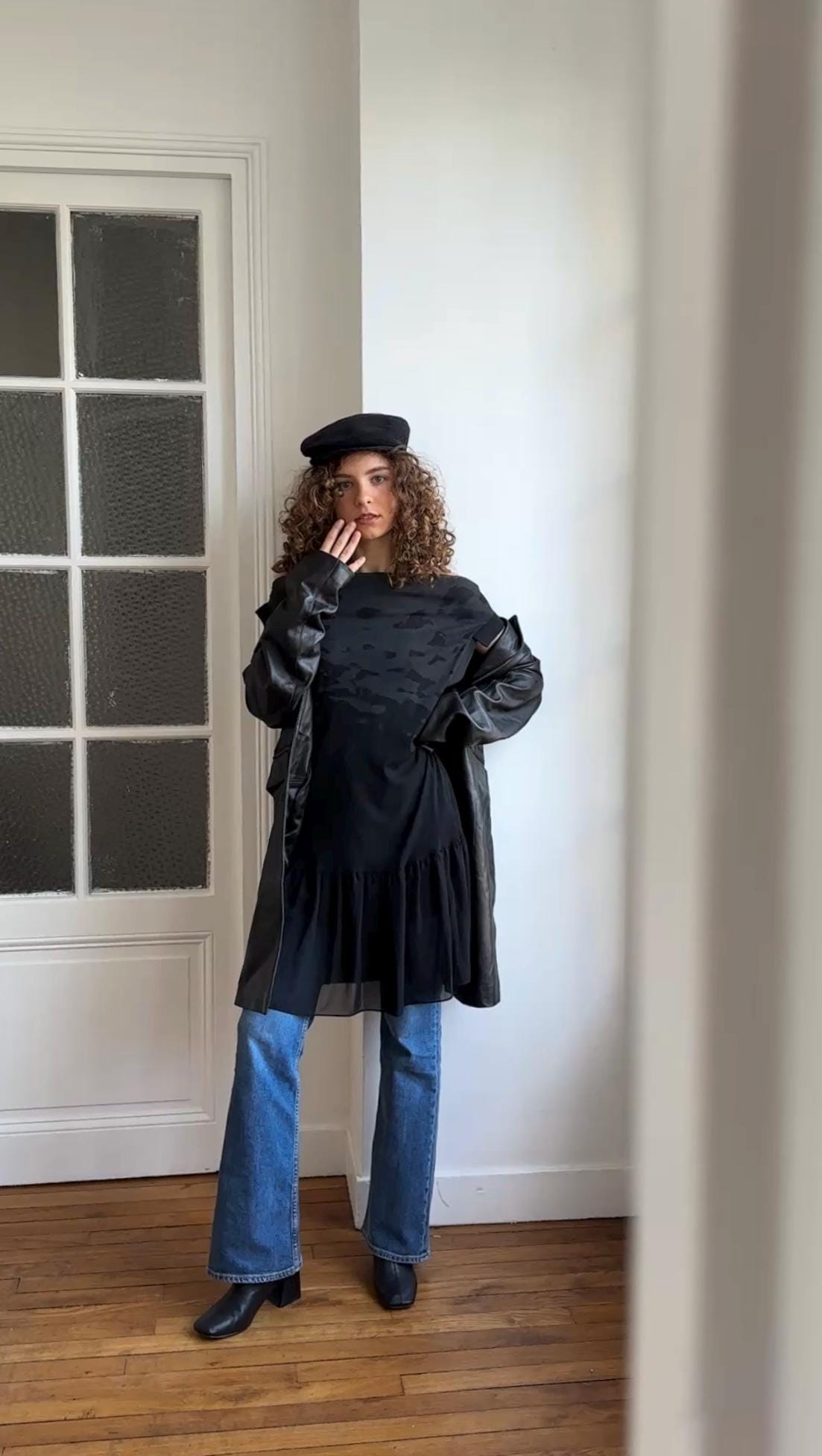 A woman with curly hair wearing a black coat, hat, and boots stands in a room with a white door and window.