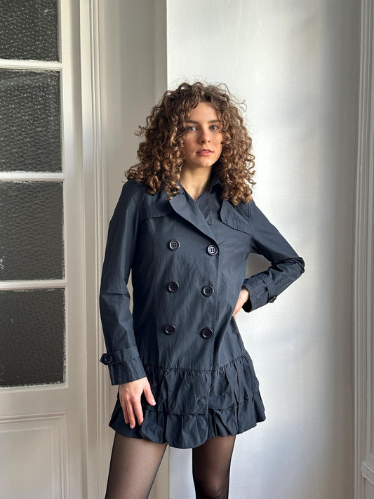 A woman with curly hair wearing a gray coat and skirt is posing in a room with a window.
