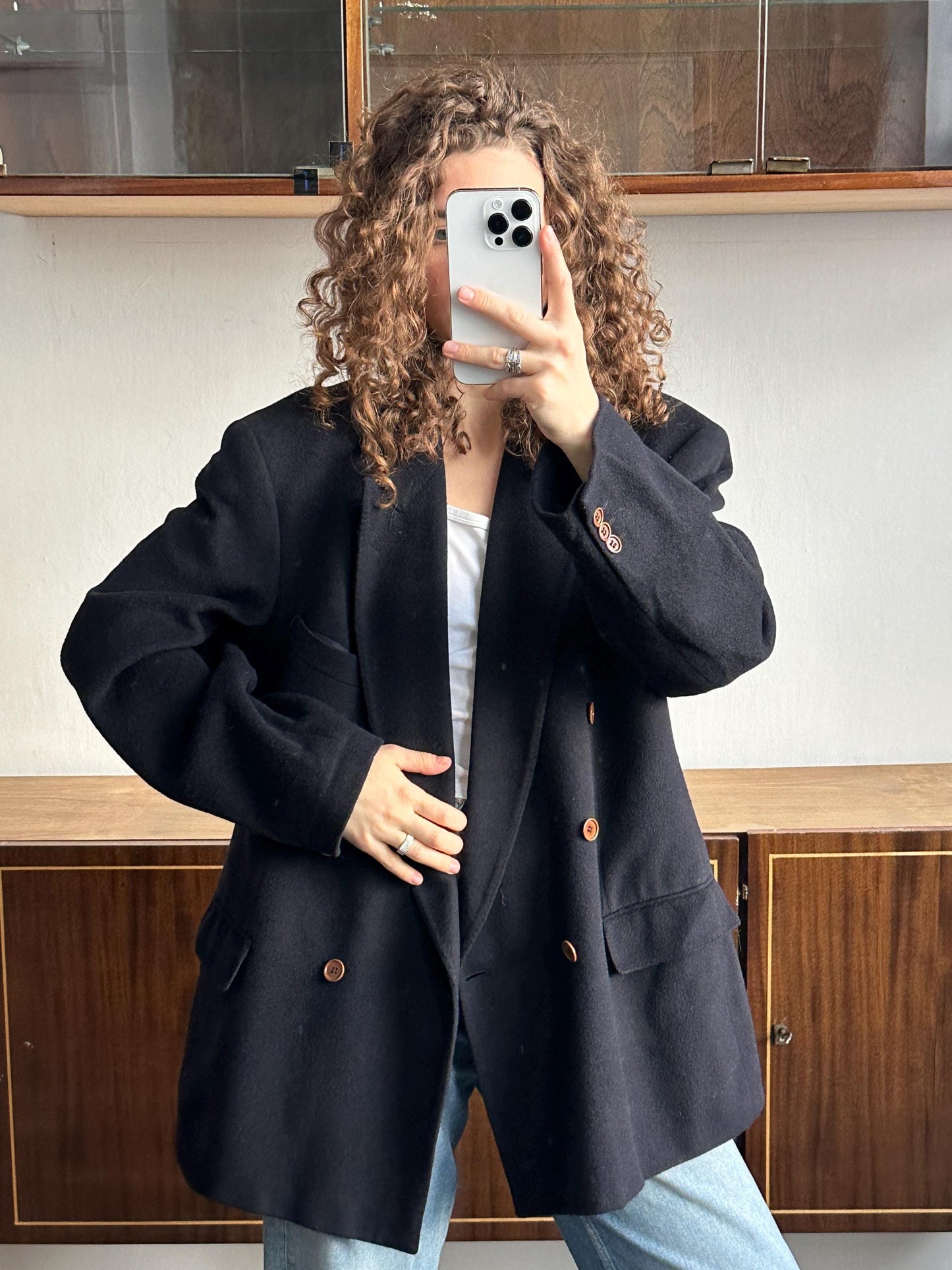A person with curly hair is taking a selfie with a white iPhone, wearing a black blazer and jeans.