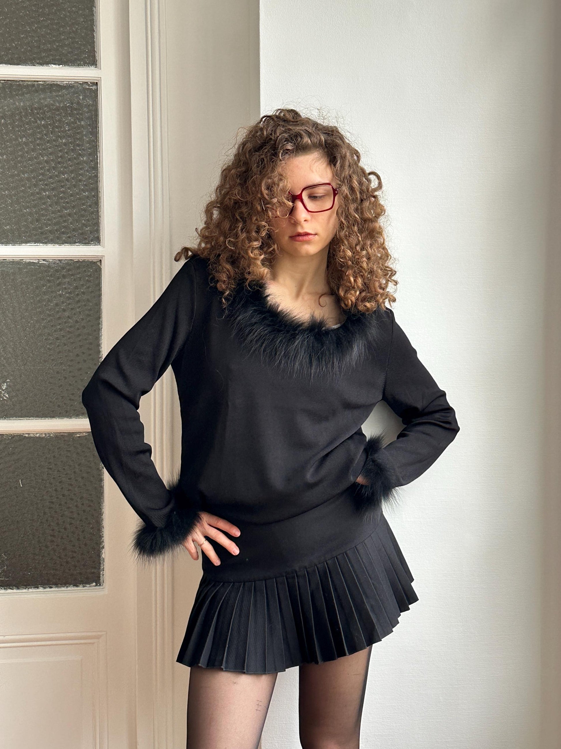 A woman with curly hair and glasses is standing in front of a white door, wearing a black outfit with a fur collar and a pleated skirt.