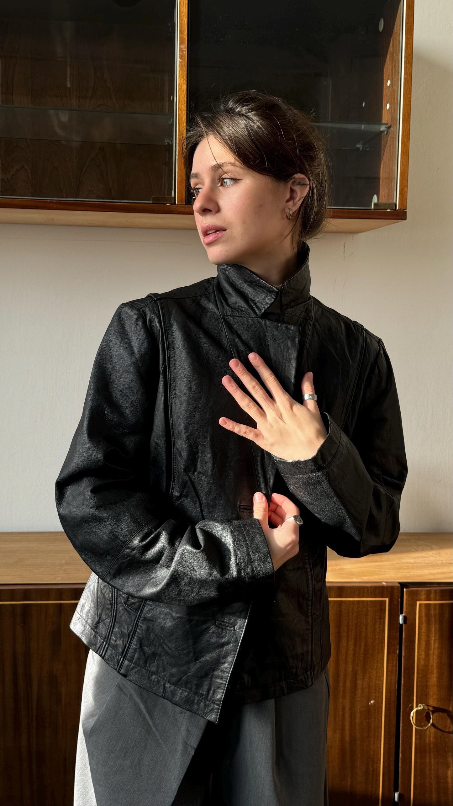 A woman in a black leather jacket stands in front of a wooden cabinet, her hand raised to her chest.