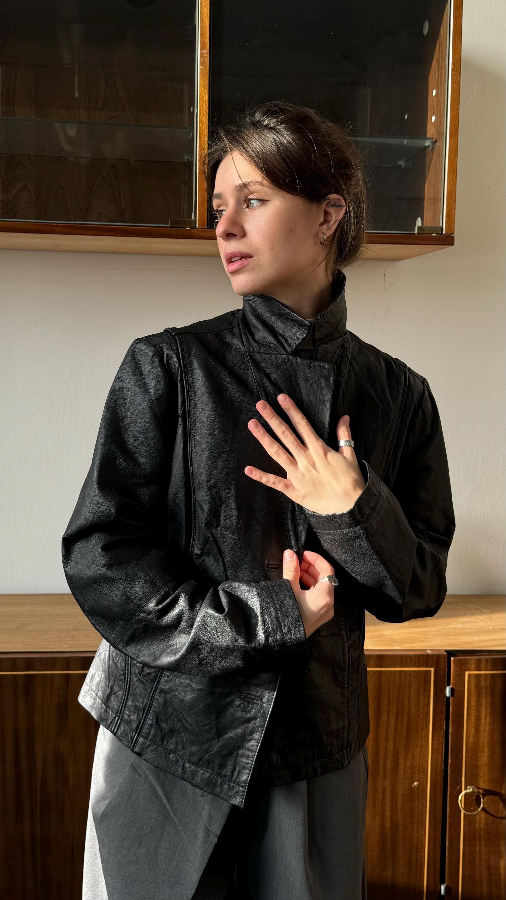 A woman in a black leather jacket stands in front of a wooden cabinet, her hand raised to her chest.