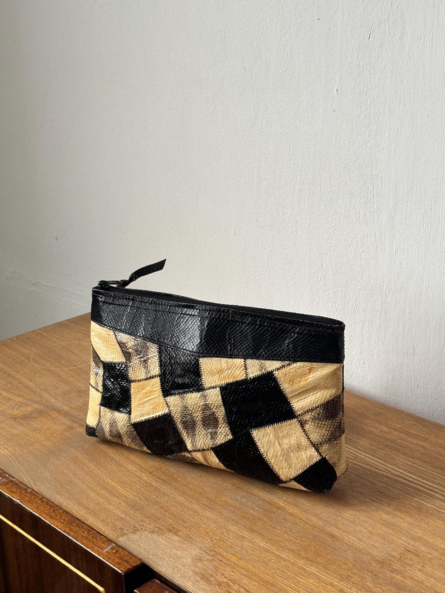 A black and tan patterned clutch purse is sitting on a wooden surface against a white wall.