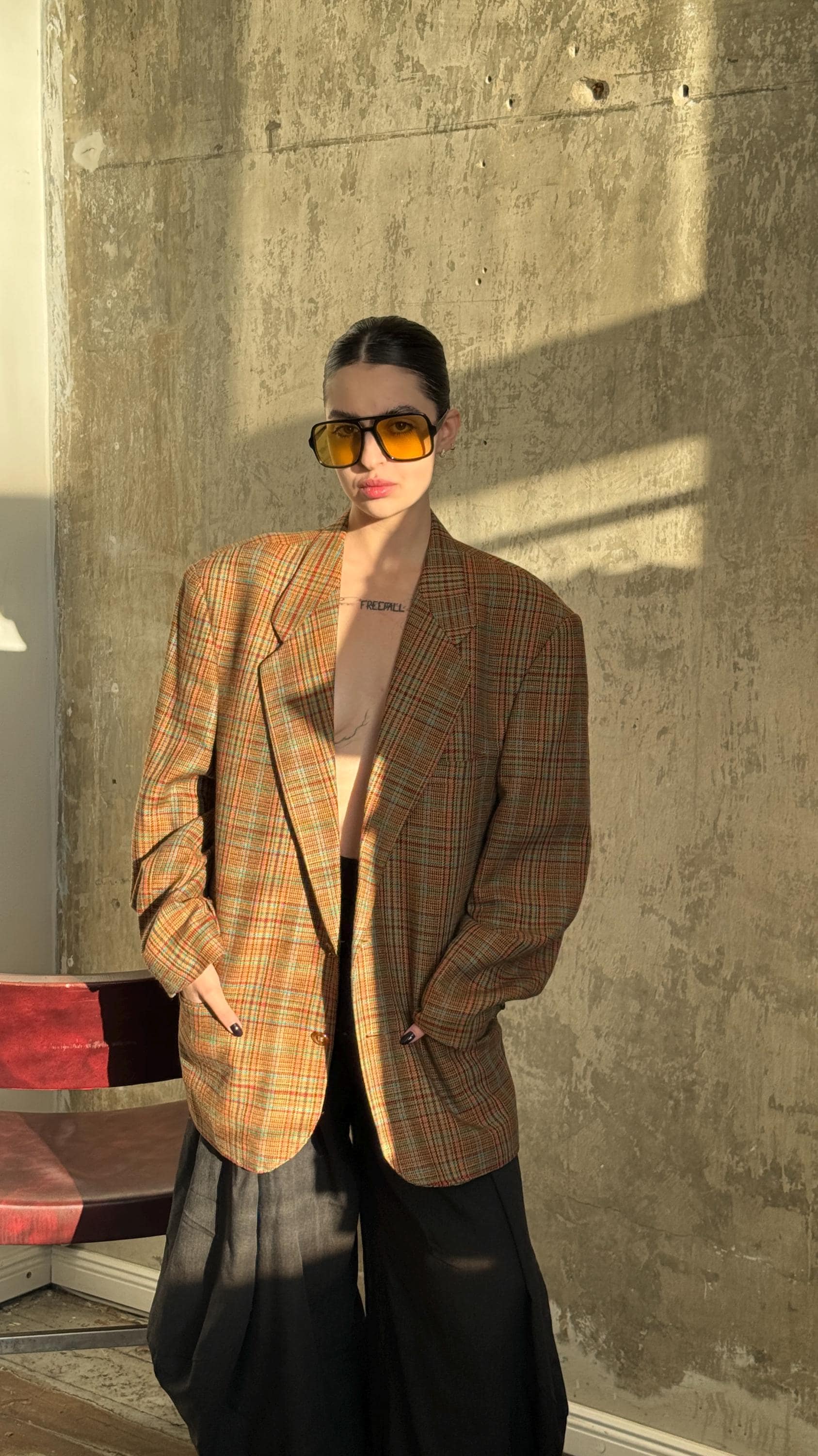 A person wearing a plaid jacket, sunglasses, and standing in front of a concrete wall.