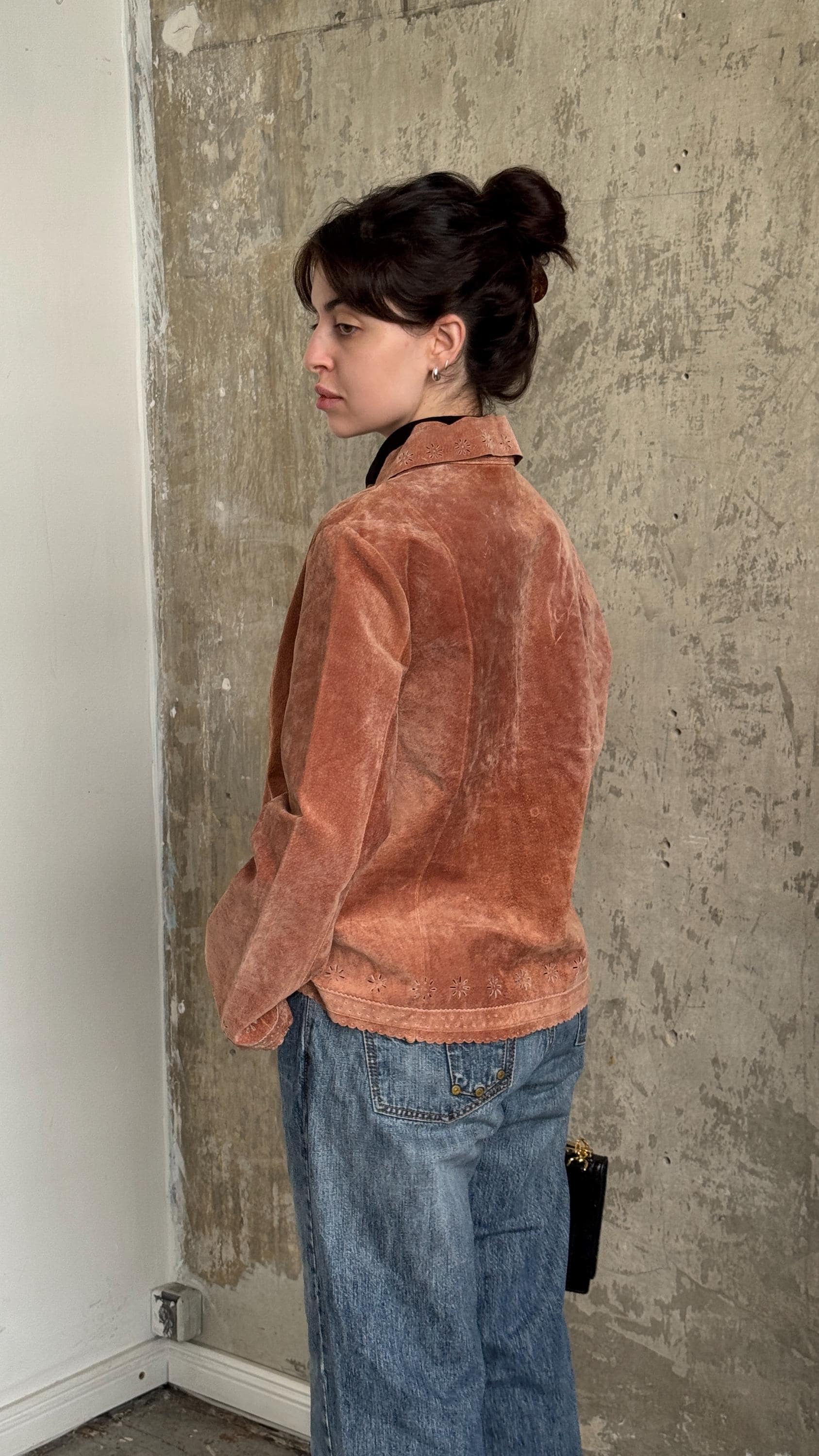 A woman wearing a brown jacket and blue jeans stands in front of a concrete wall.