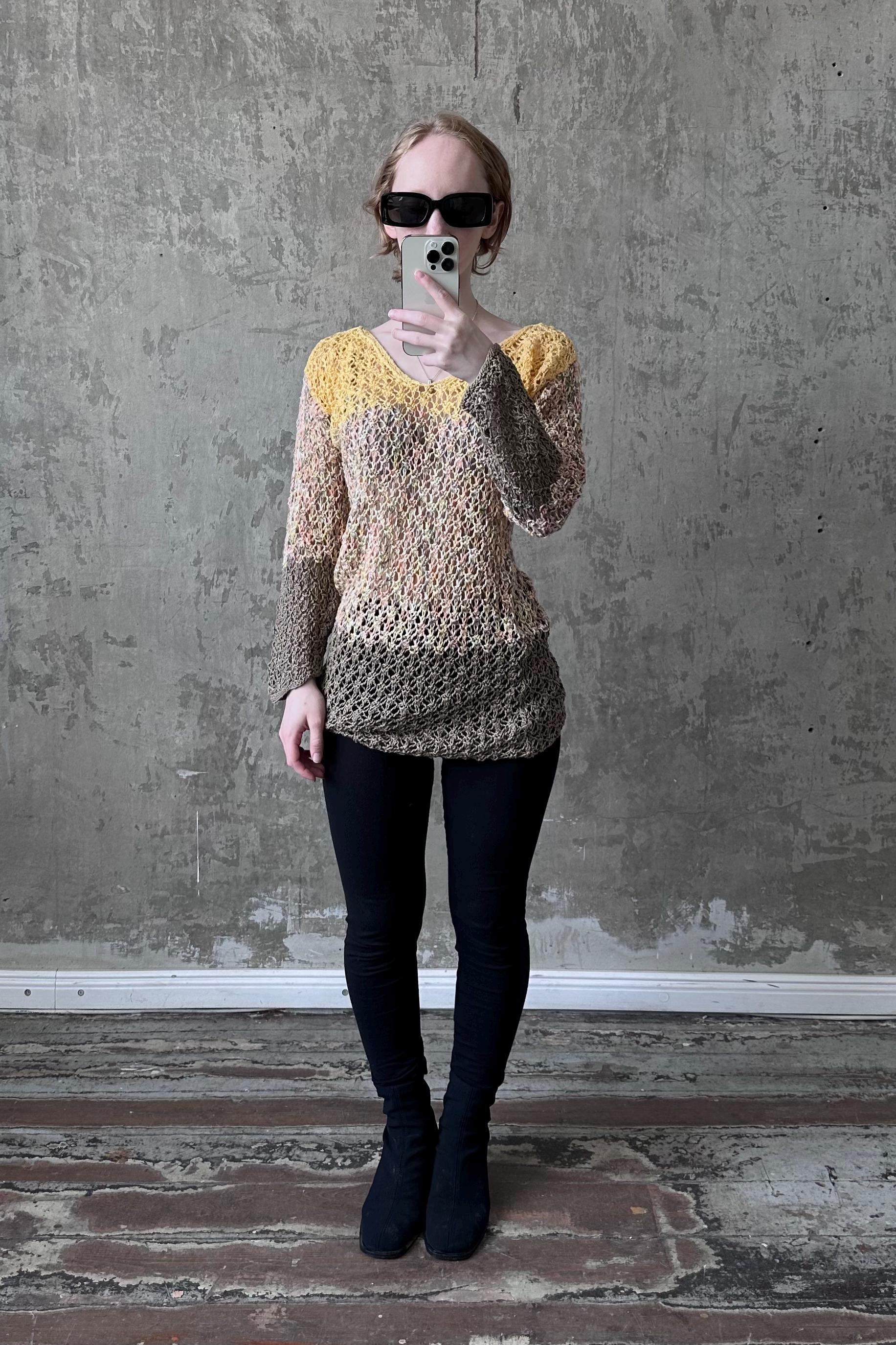 A woman wearing a yellow and brown patterned top, black leggings, and black boots is taking a selfie with a smartphone against a concrete wall.