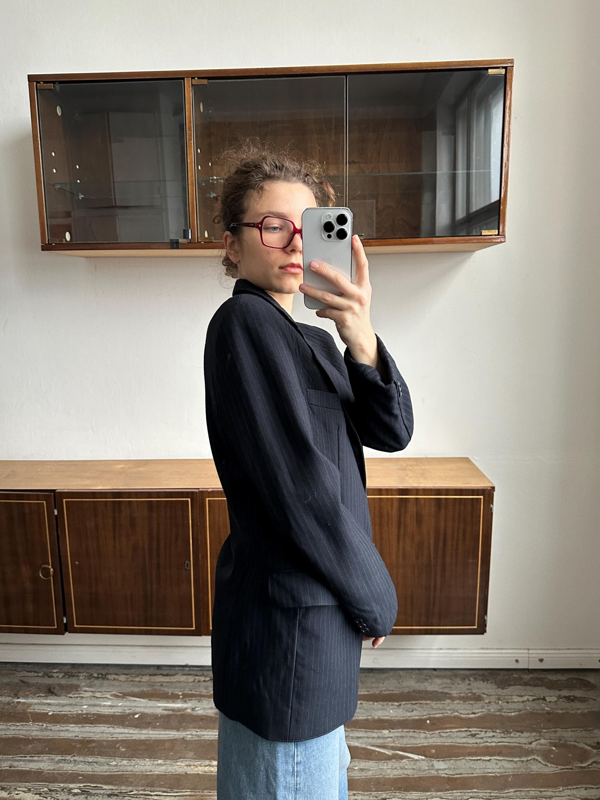 A woman wearing a black blazer and red glasses is taking a selfie with her iPhone in front of a wooden cabinet.