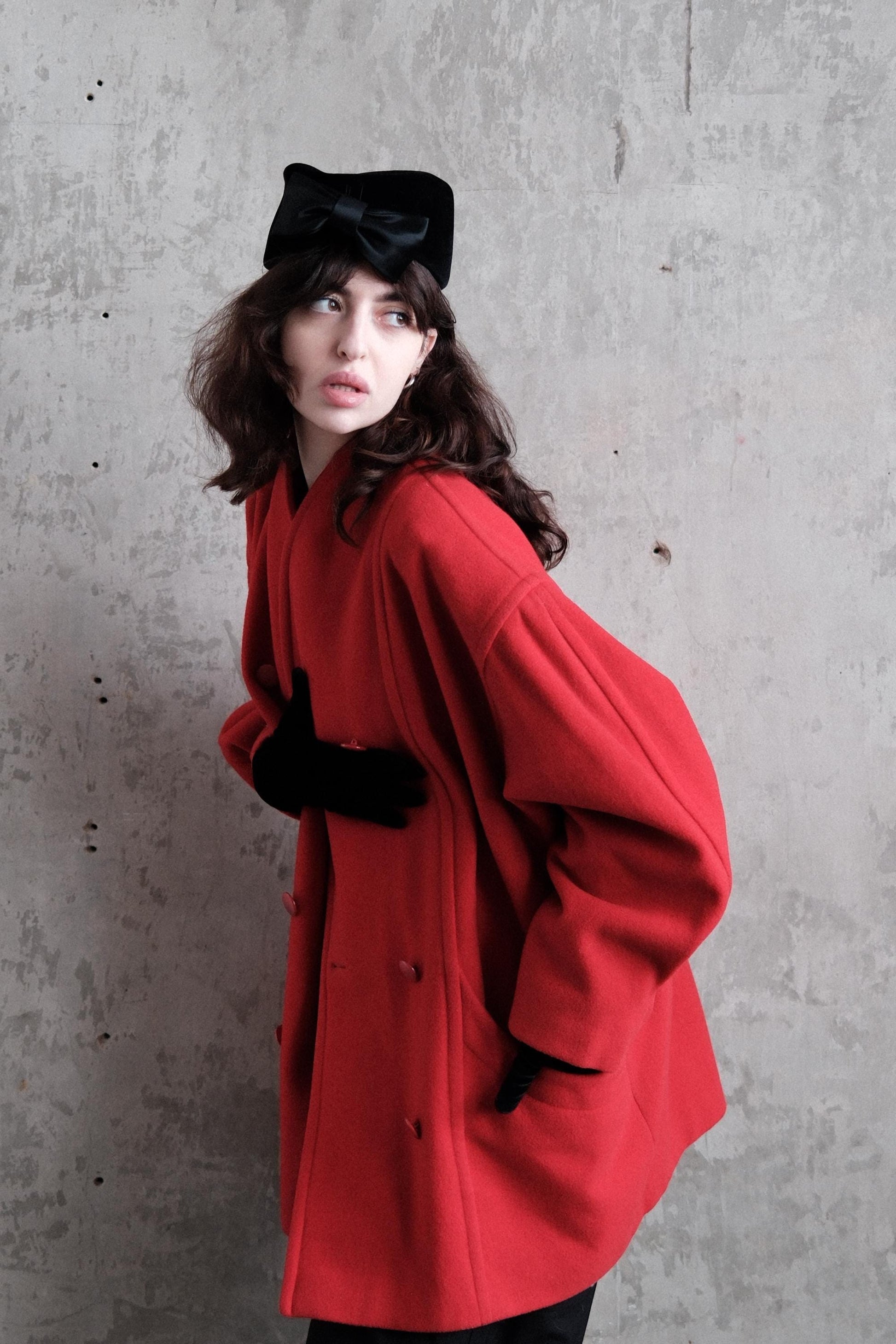 A woman wearing a red coat and a black hat is posing against a concrete wall.