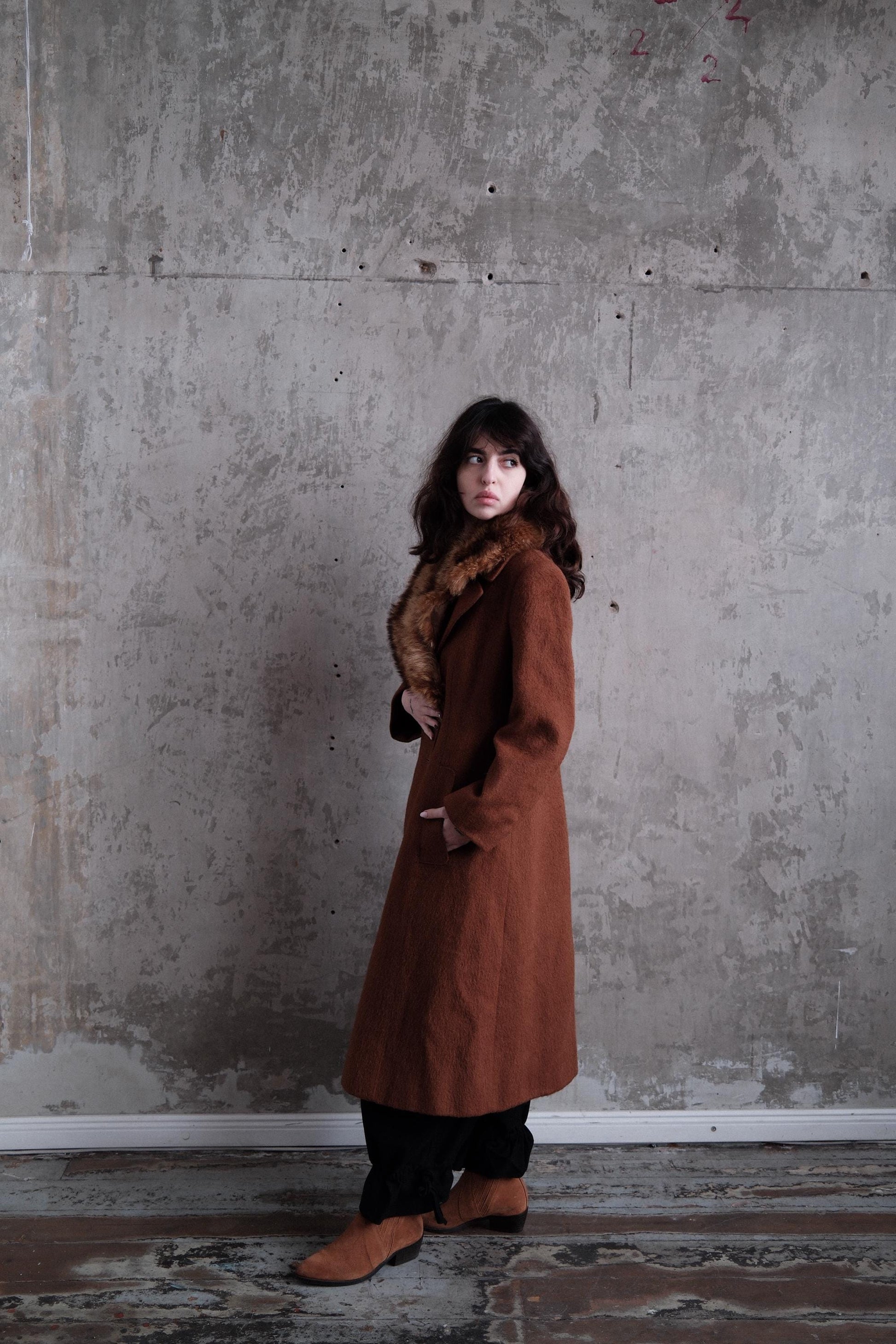A woman in a long brown coat stands in a concrete room, her gaze directed off to the side.