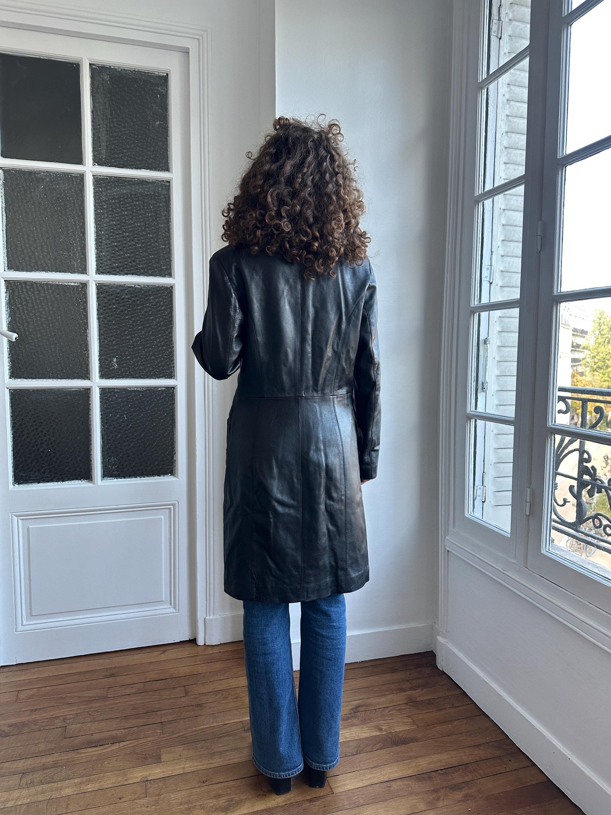 A person with curly hair is standing in a room, facing away from the camera, wearing a black leather coat.