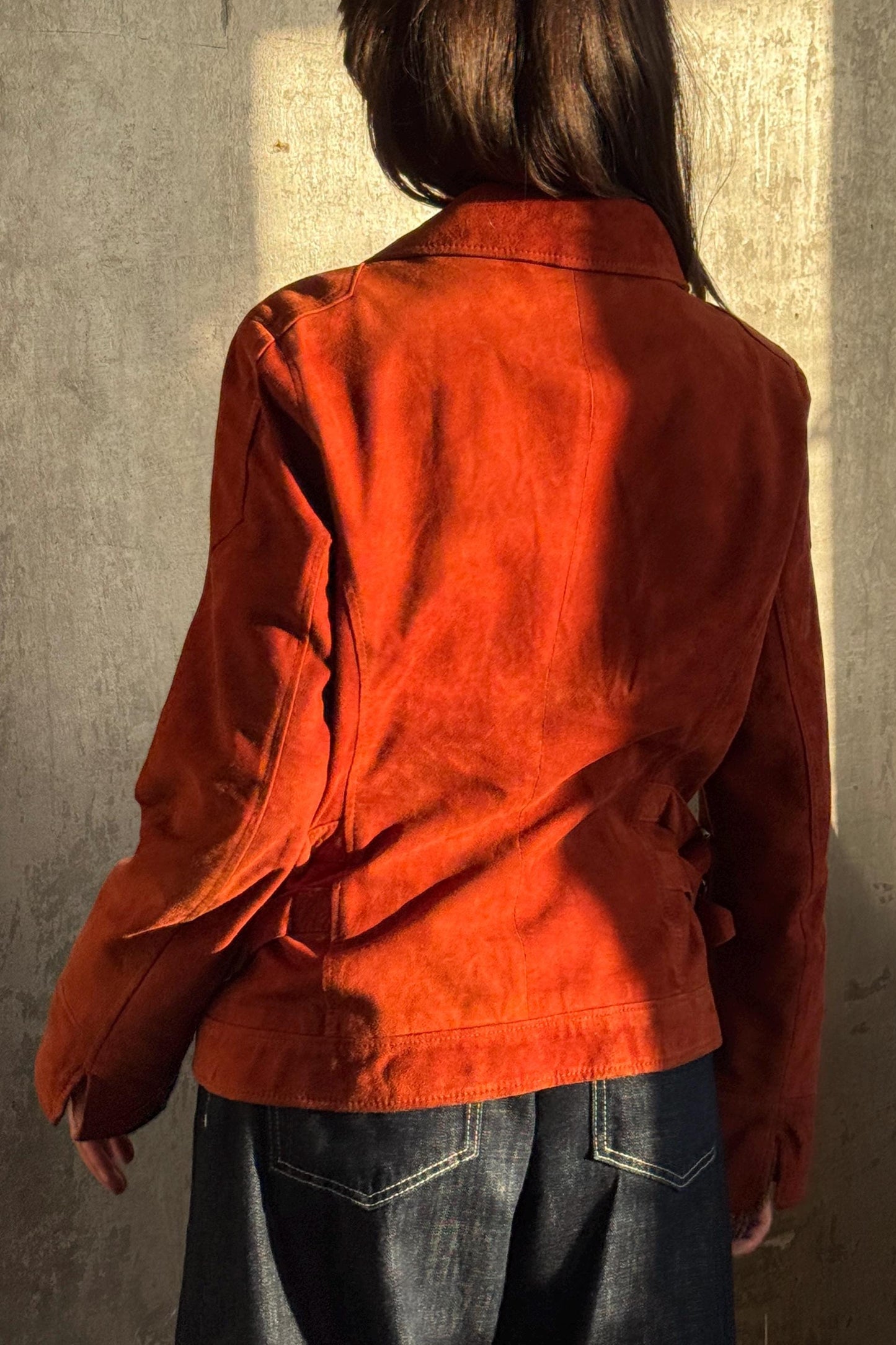 the back of a person wearing a red jacket and blue jeans, standing against a concrete wall.
