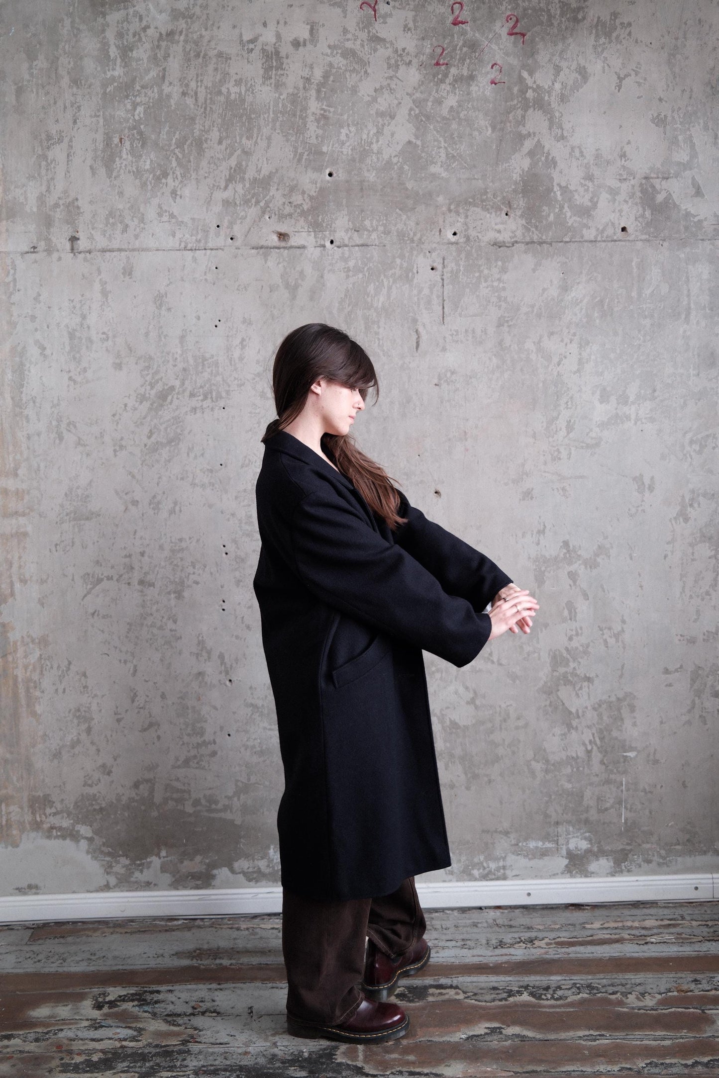 A person in a long black coat stands against a concrete wall, their head tilted downwards.