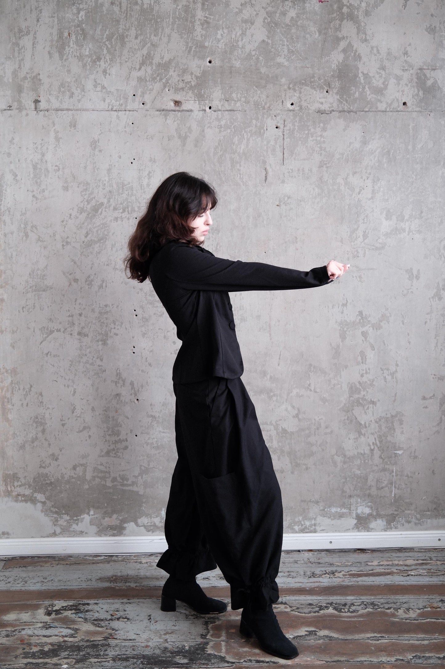 A woman in a black outfit stands in a room with a concrete wall, her head tilted downwards.