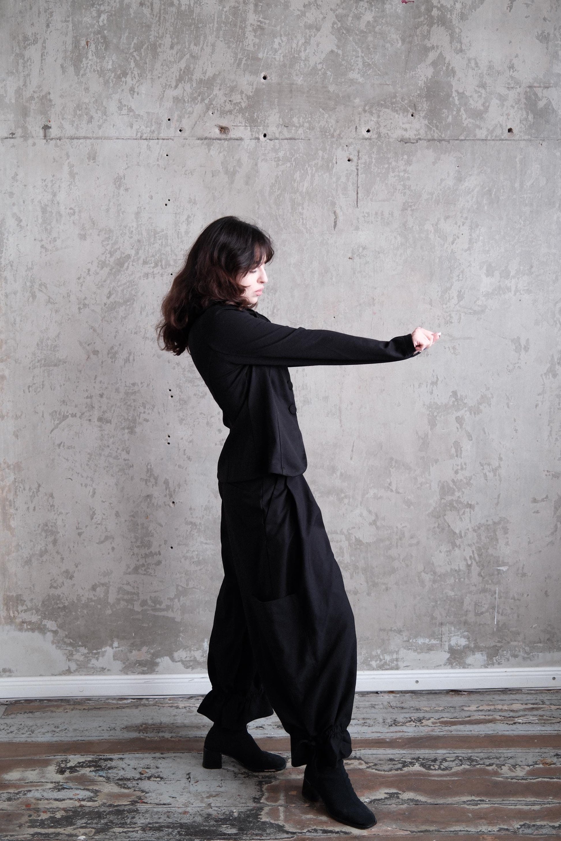 A woman in a black outfit stands in a room with a concrete wall, her head tilted downwards.