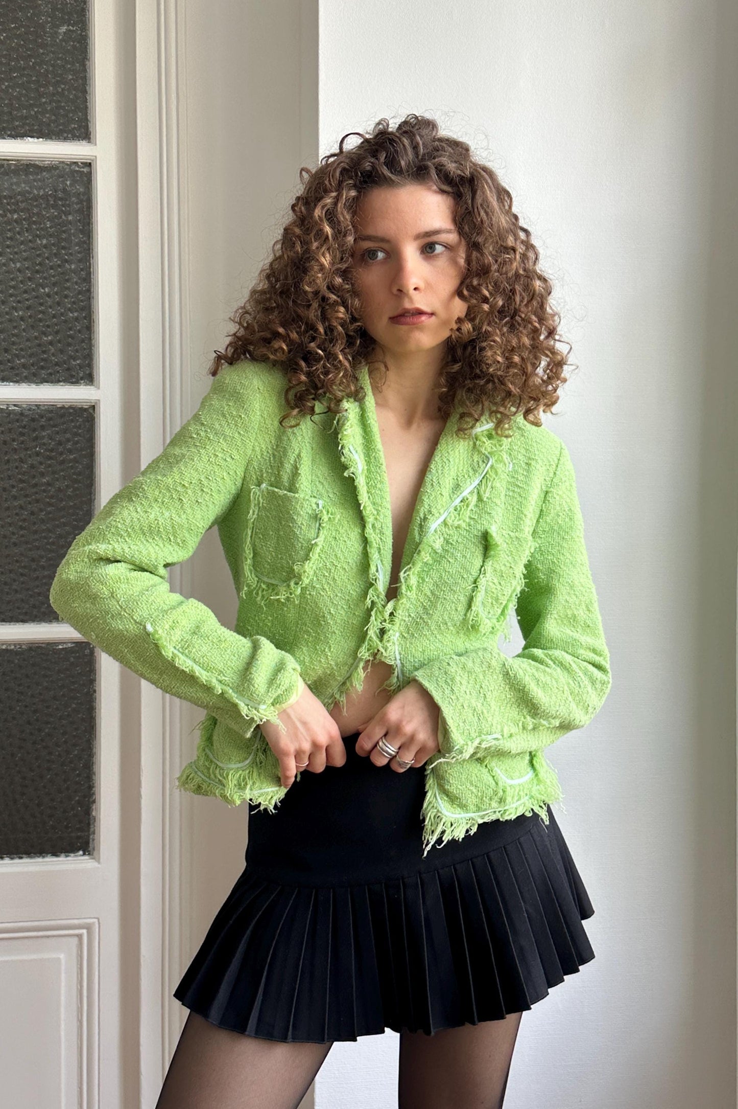 A woman with curly hair wearing a lime green jacket and a black pleated skirt stands in front of a white door.