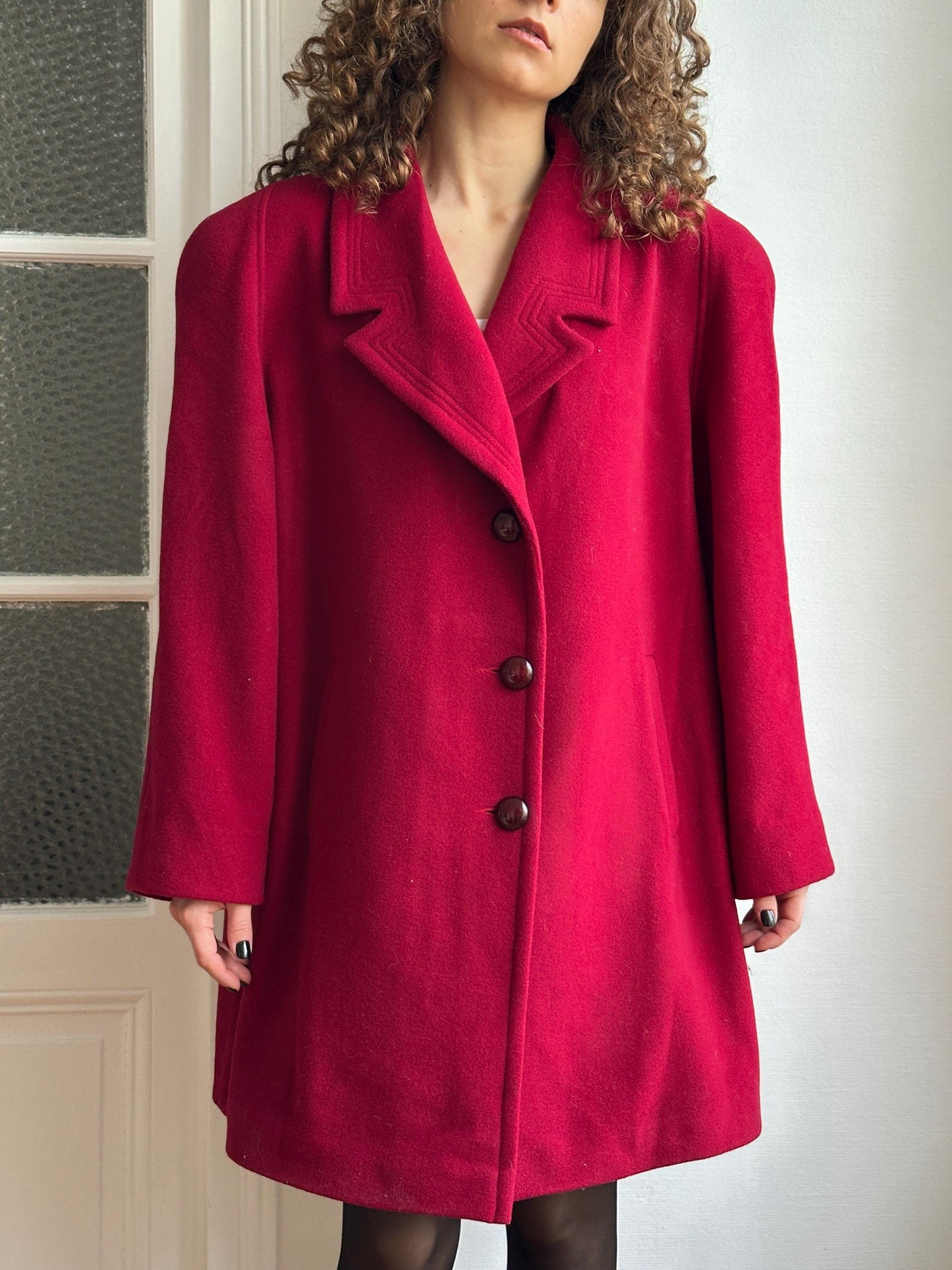 A person wearing a red coat with three buttons, standing against a white wall.